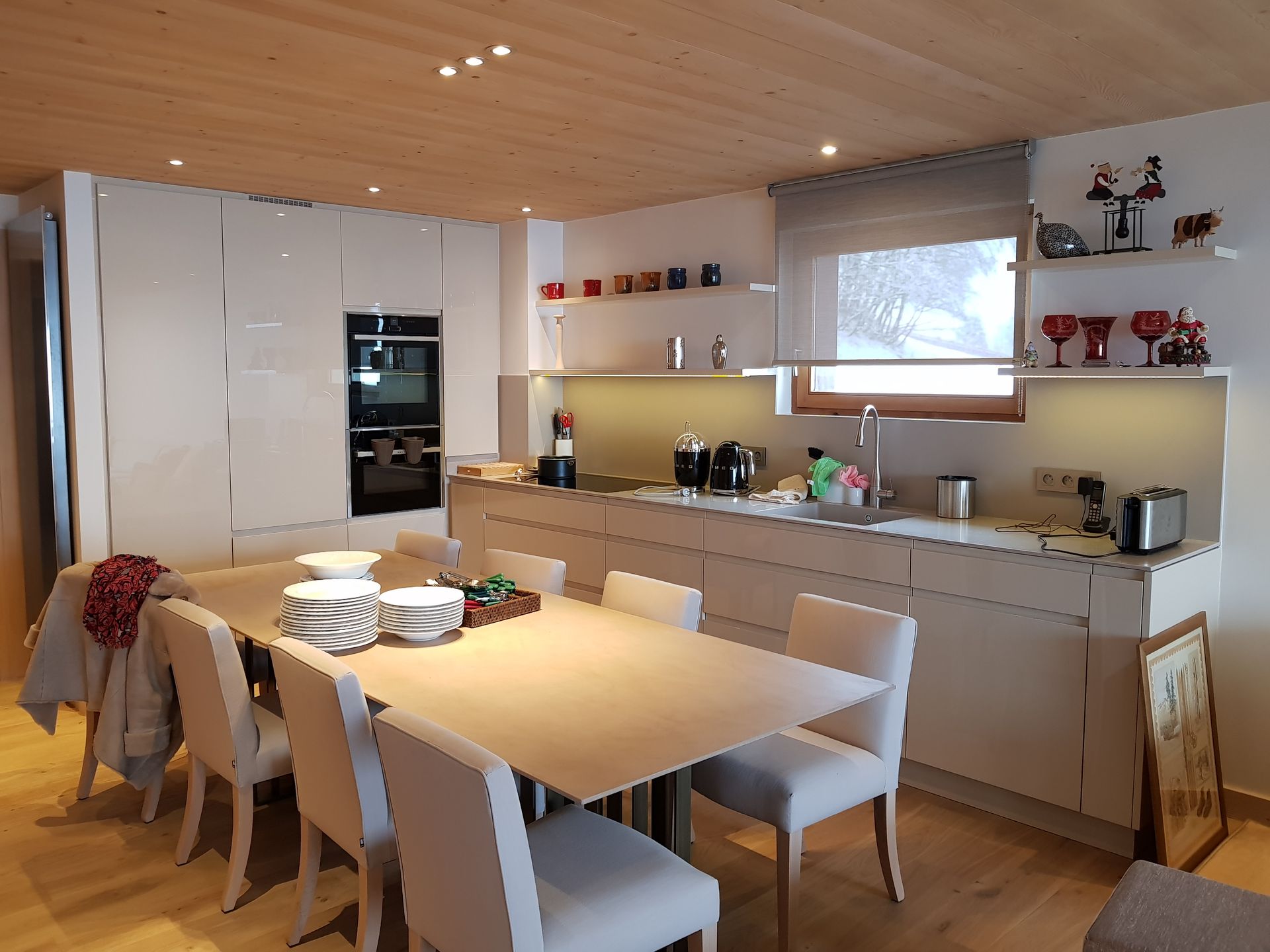 Cuisine et salle à manger modernes avec plafond en bois clair, placards blancs et une longue table dressée pour les repas.
