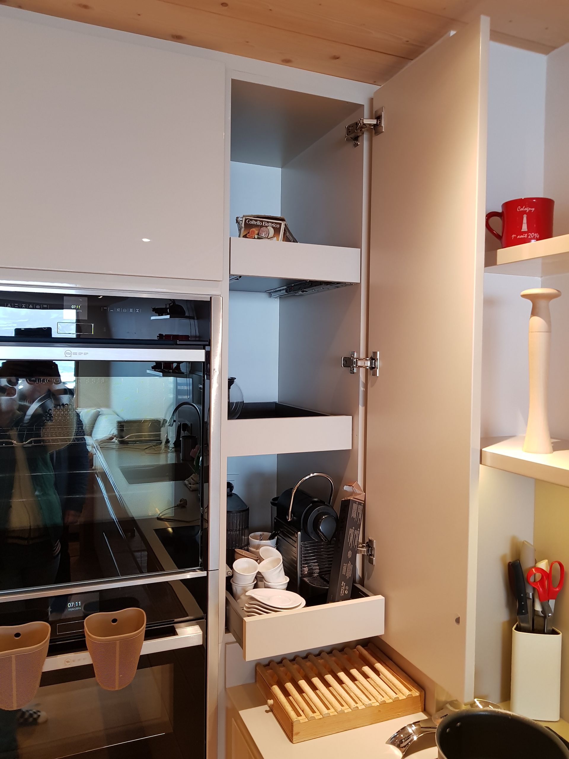 Meuble de cuisine blanc avec étagères coulissantes contenant une machine à café et des tasses, à côté d'un four encastré.