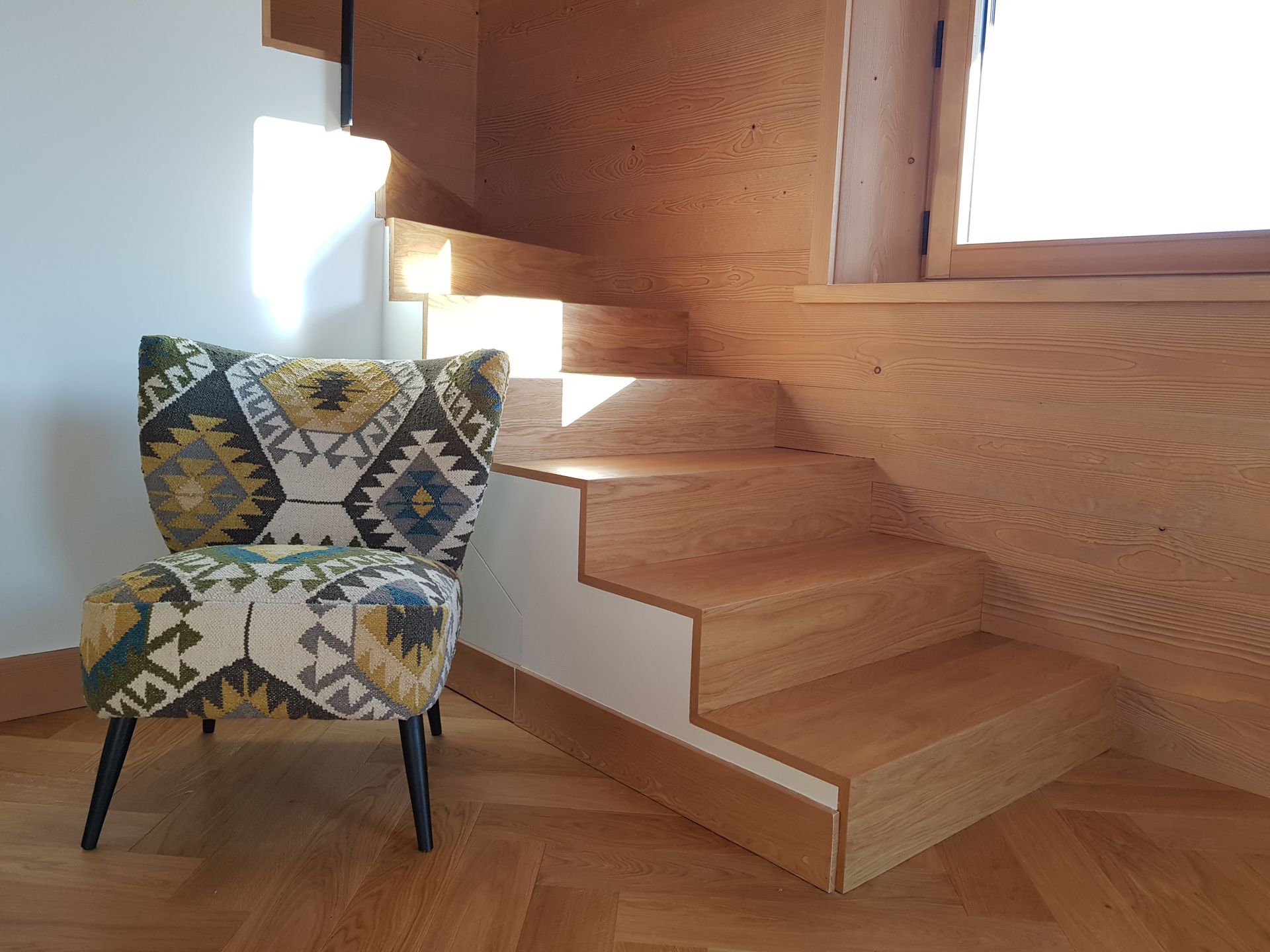 Fauteuil recouvert d'un tissu à motifs, placé à côté d'un escalier en bois dans une pièce baignée de soleil.
