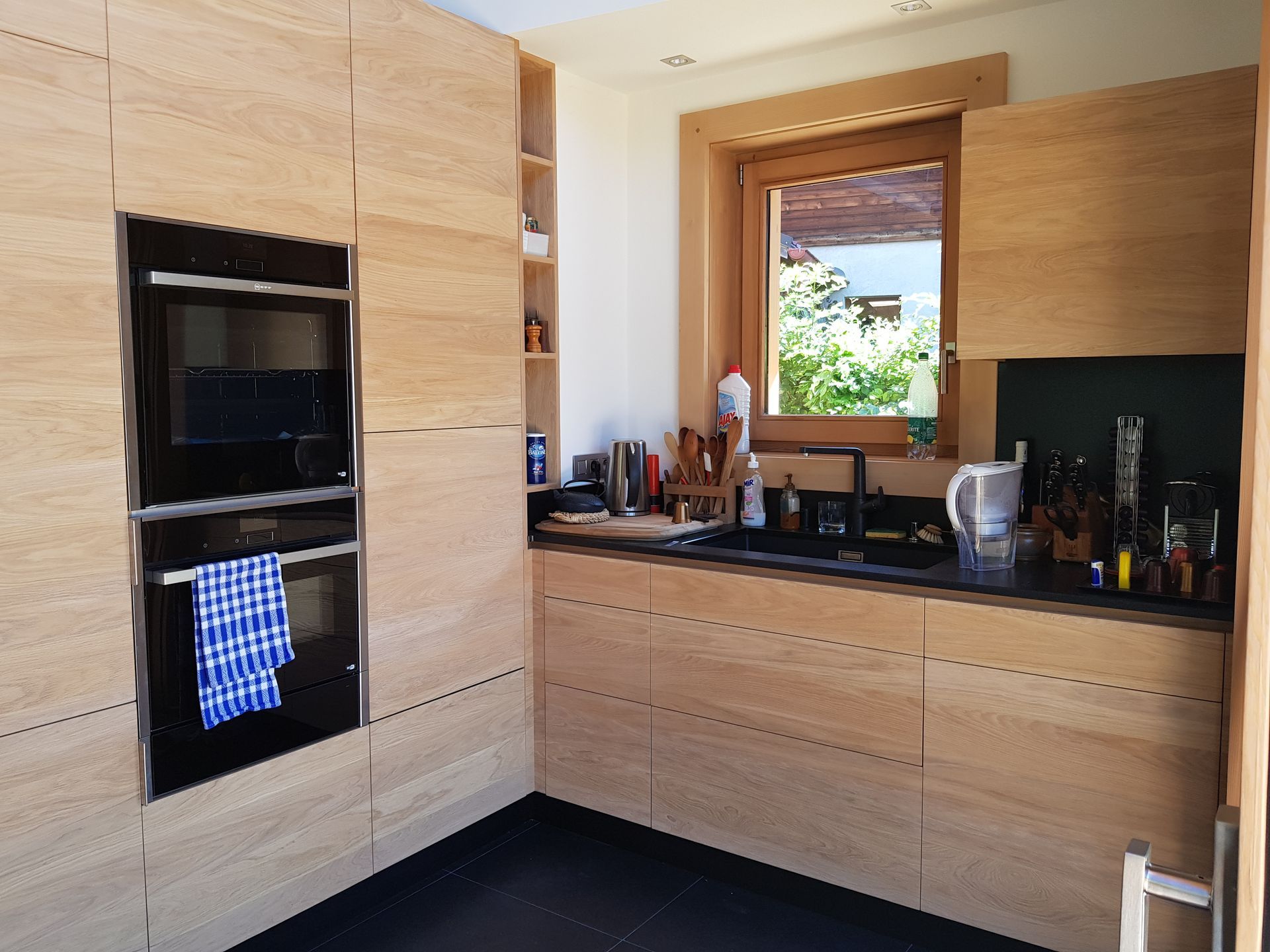 Cuisine moderne avec placards en bois clair, four encastré noir et évier près d'une fenêtre.