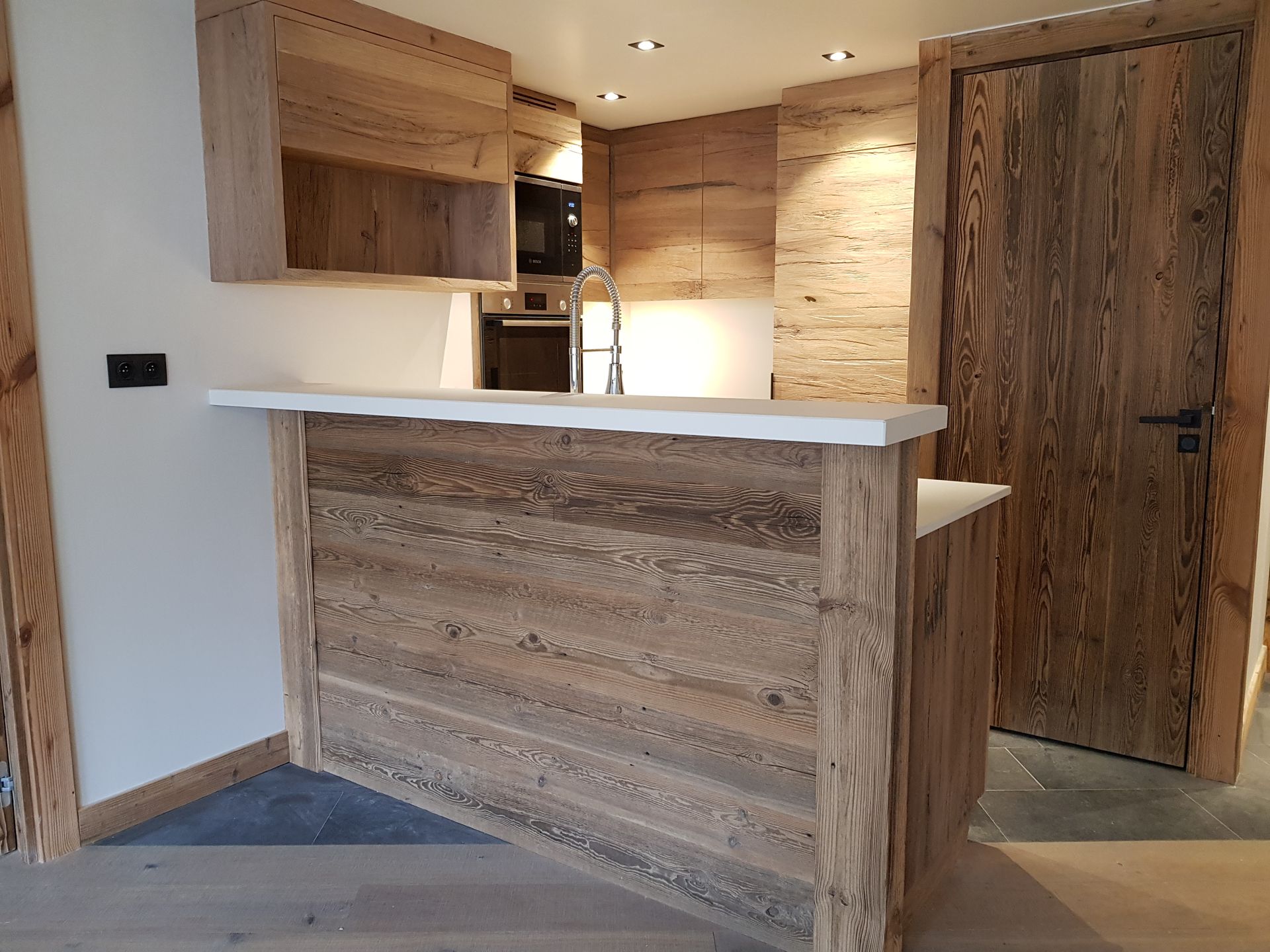 Cuisine en bois avec bar et électroménager. La porte, les placards et le bar sont en bois. Plan de travail blanc.