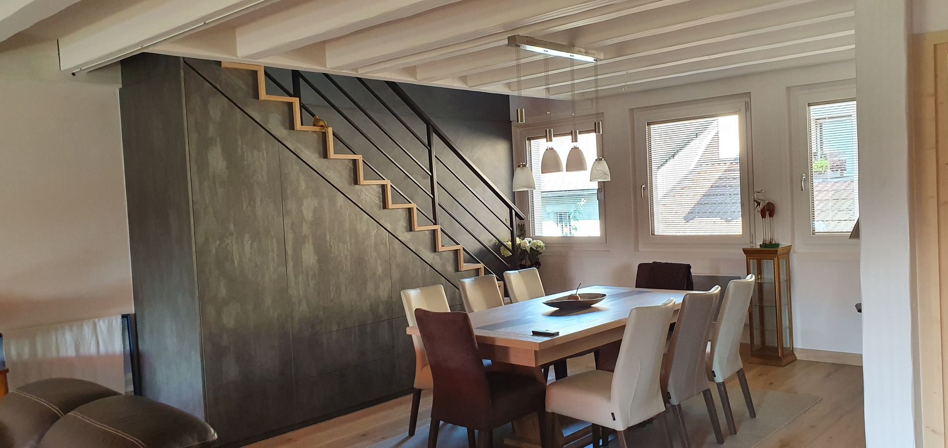 Salle à manger avec escalier, plafond à poutres apparentes et grande table avec chaises.