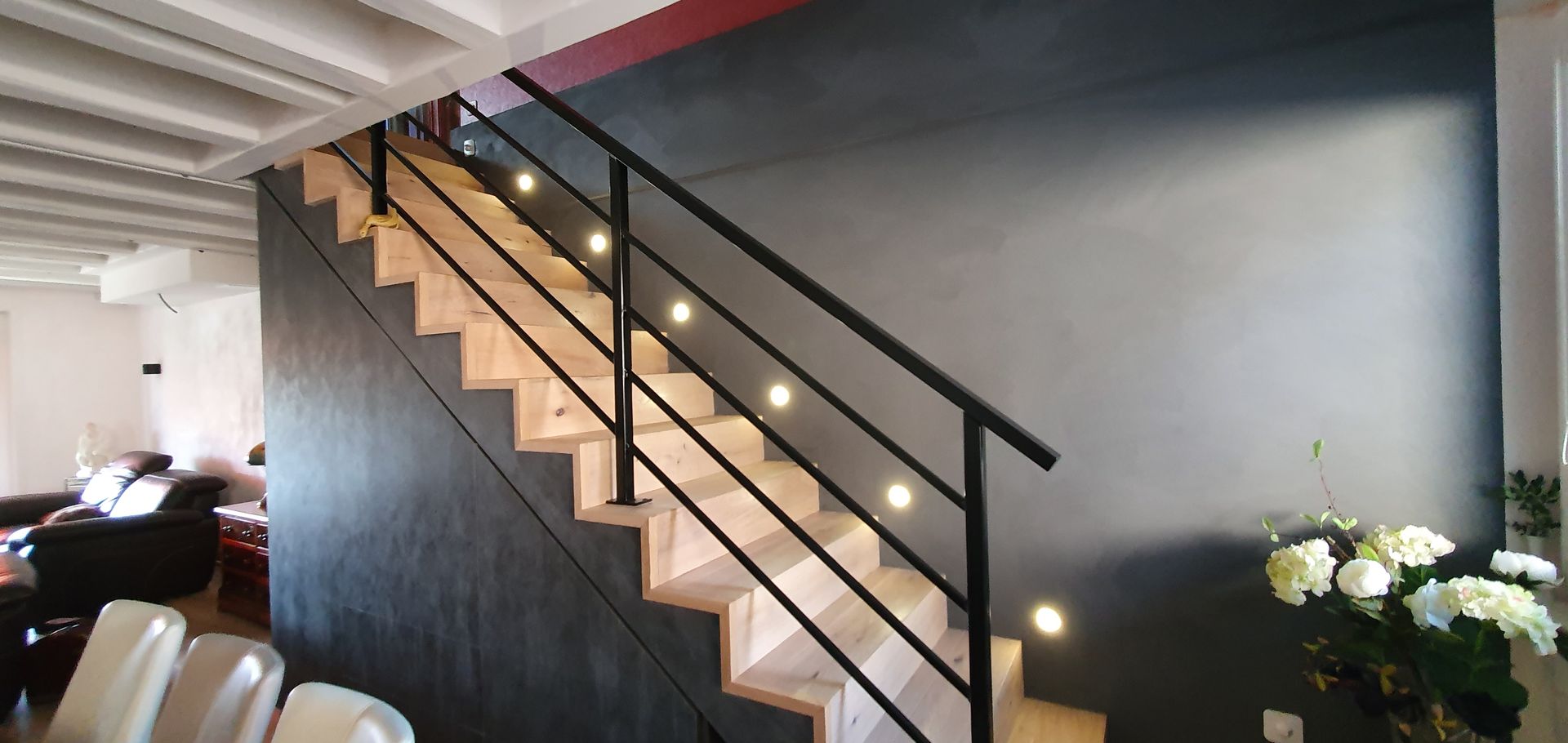 Escalier en bois avec rampe en métal noir et éclairage intégré, adossé à un mur gris foncé.