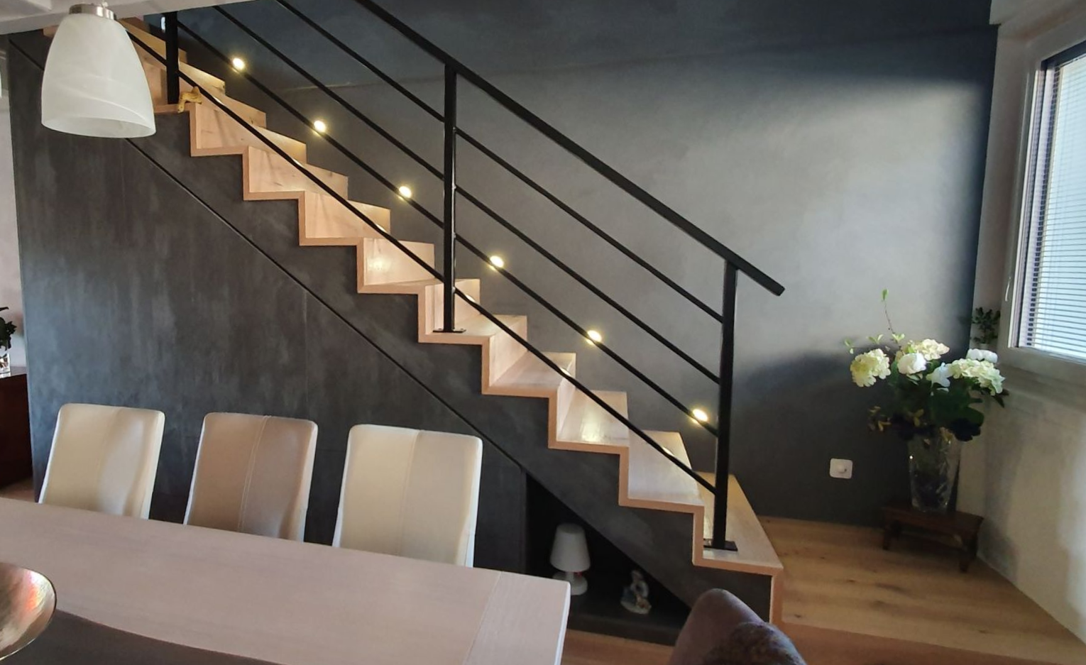 Escalier en bois avec rampe en métal noir, éclairé par de petites lumières, dans un intérieur moderne.