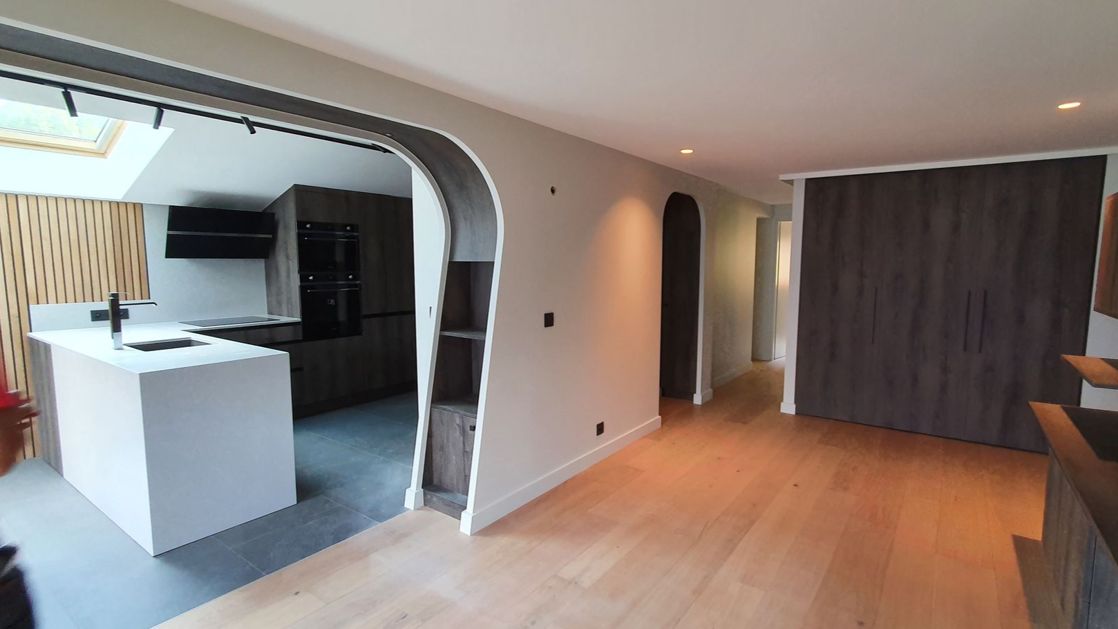 Cuisine et séjour à aire ouverte avec arcades, parquet clair et murs aux tons neutres. La cuisine comprend un îlot blanc et des appareils électroménagers foncés. 