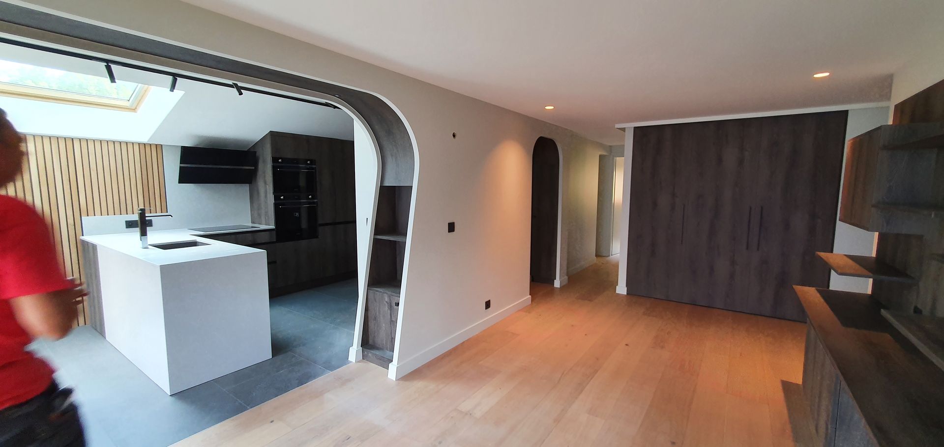 Un espace de vie ouvert avec une cuisine visible à travers une arche. On y trouve notamment un parquet et des placards en bois foncé.