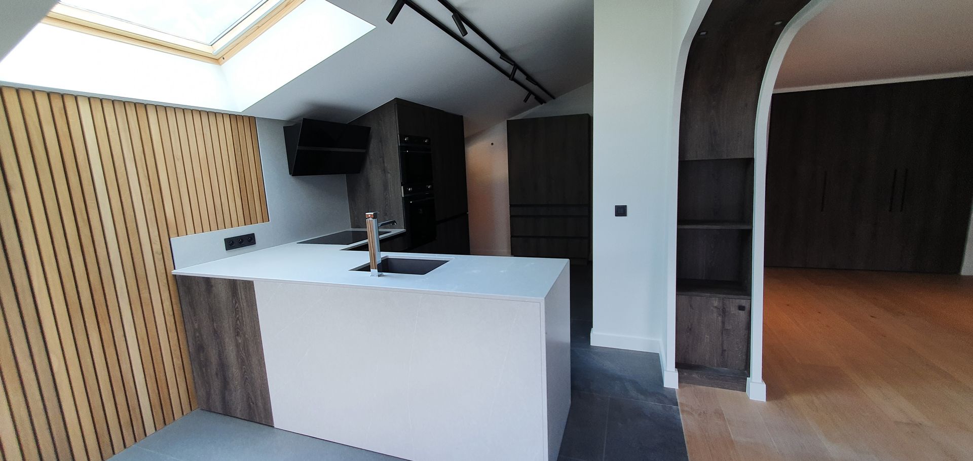 Cuisine moderne avec un mur d'accent en bois, un îlot blanc et une arche menant à une pièce avec parquet.