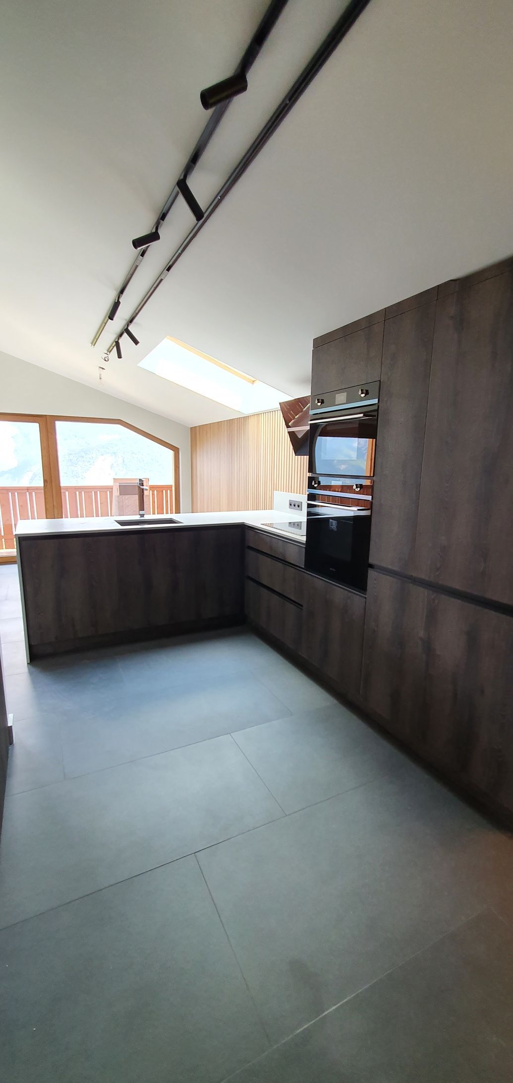 Cuisine moderne avec des armoires en bois foncé, un grand îlot central et un plafond mansardé avec un puits de lumière et des spots sur rail.