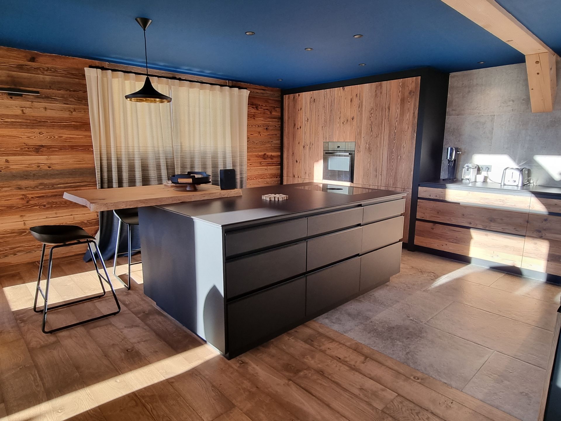 Cuisine moderne avec îlot gris, murs en bois, plafond bleu et table en bois.
