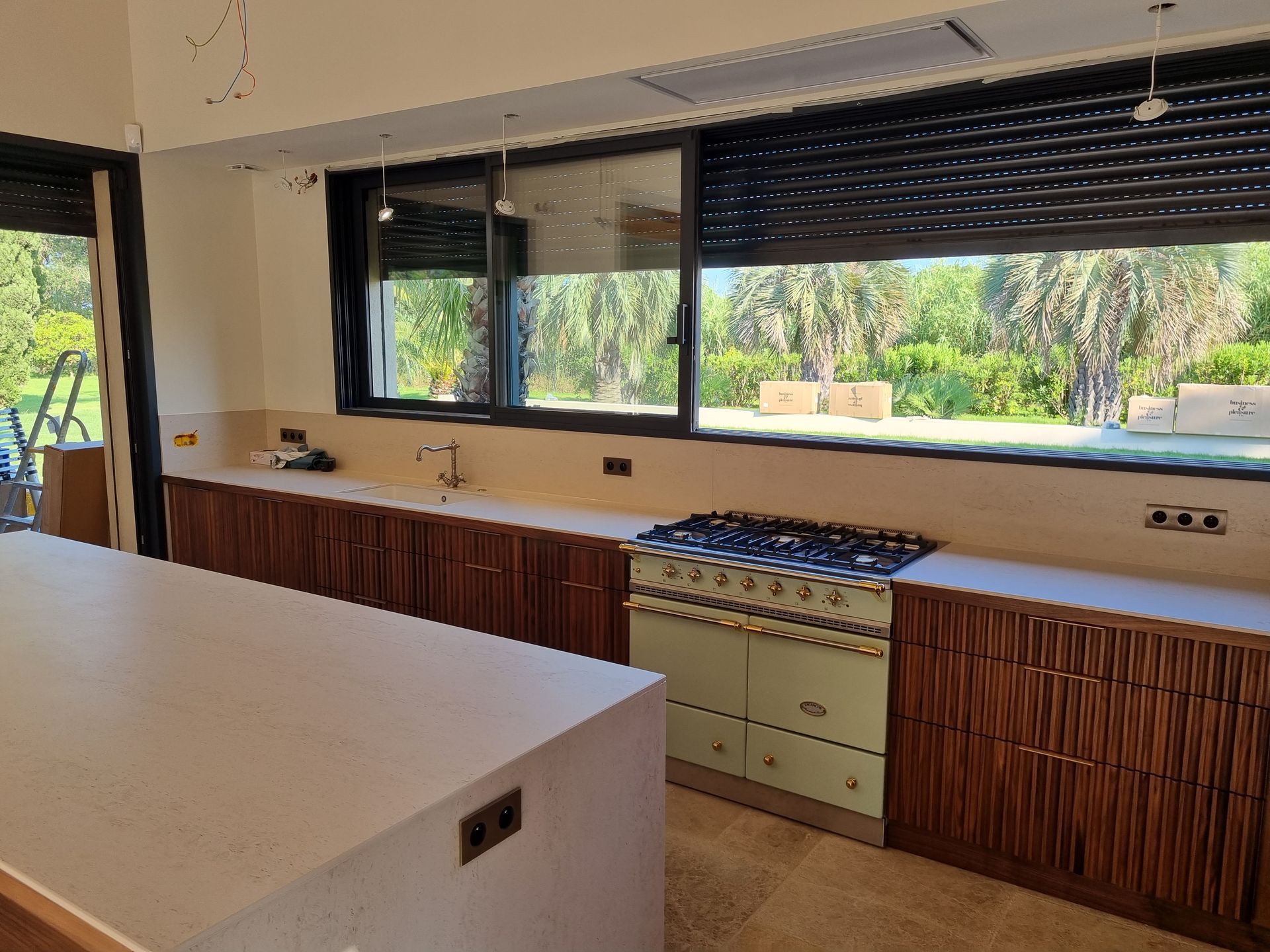 Cuisine avec plans de travail clairs, placards en bois foncé, cuisinière vert clair et grande fenêtre donnant sur la verdure.