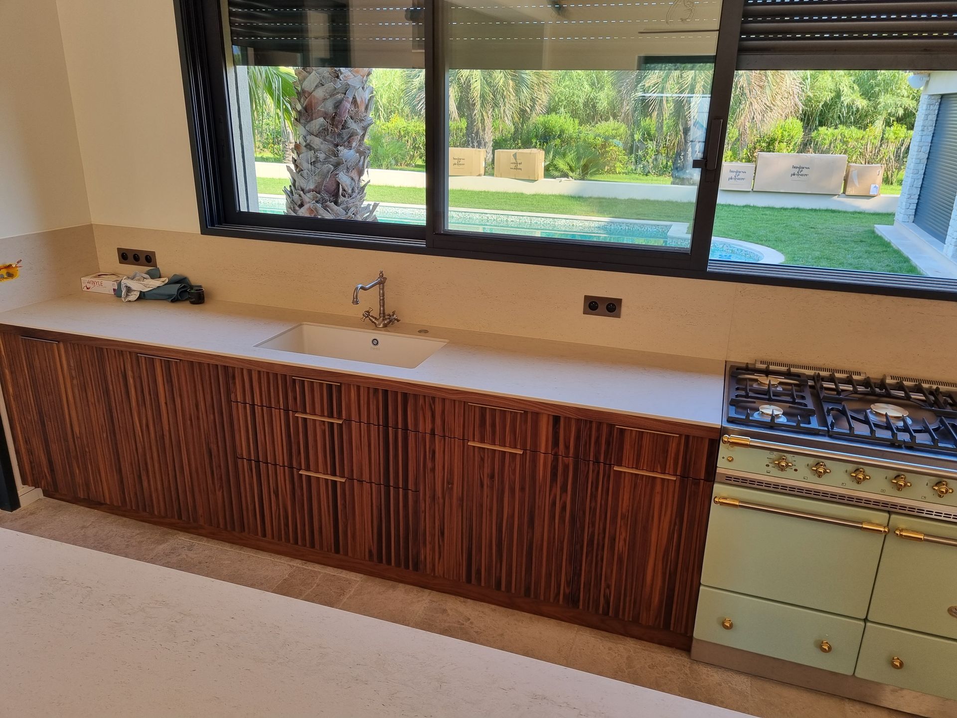 Cuisine avec placards en bois, plan de travail clair, évier et plaque de cuisson. La fenêtre donne sur un jardin verdoyant.