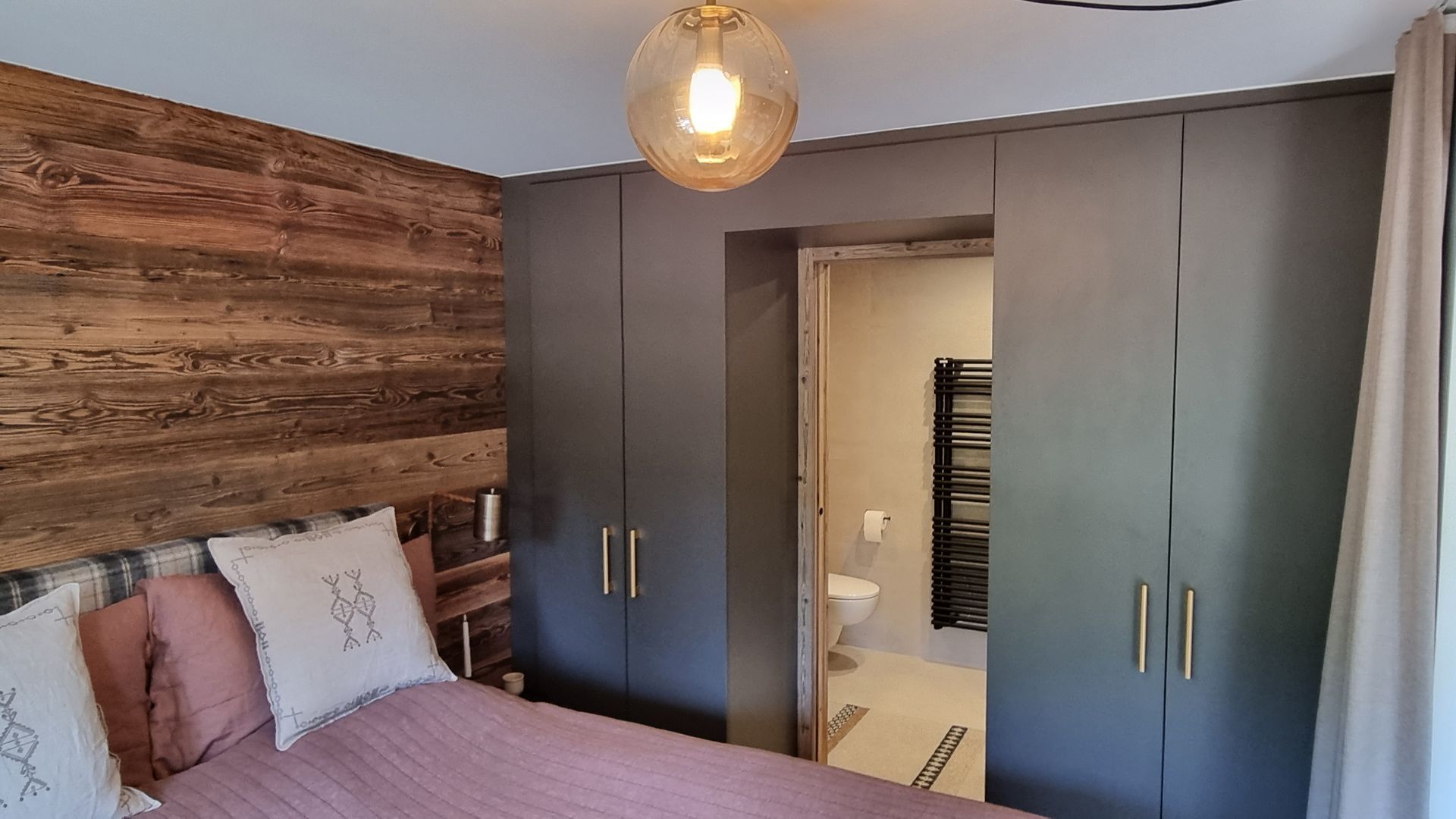 Chambre avec un mur en bois apparent, une armoire grise, un lit et une salle de bains partiellement visible.
