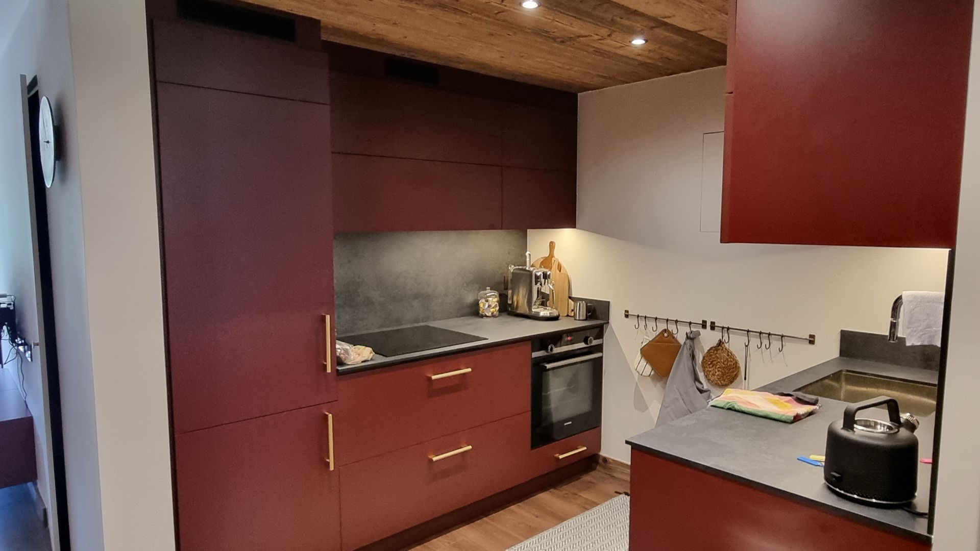 Cuisine bordeaux avec comptoirs foncés, four encastré et plafond en bois.