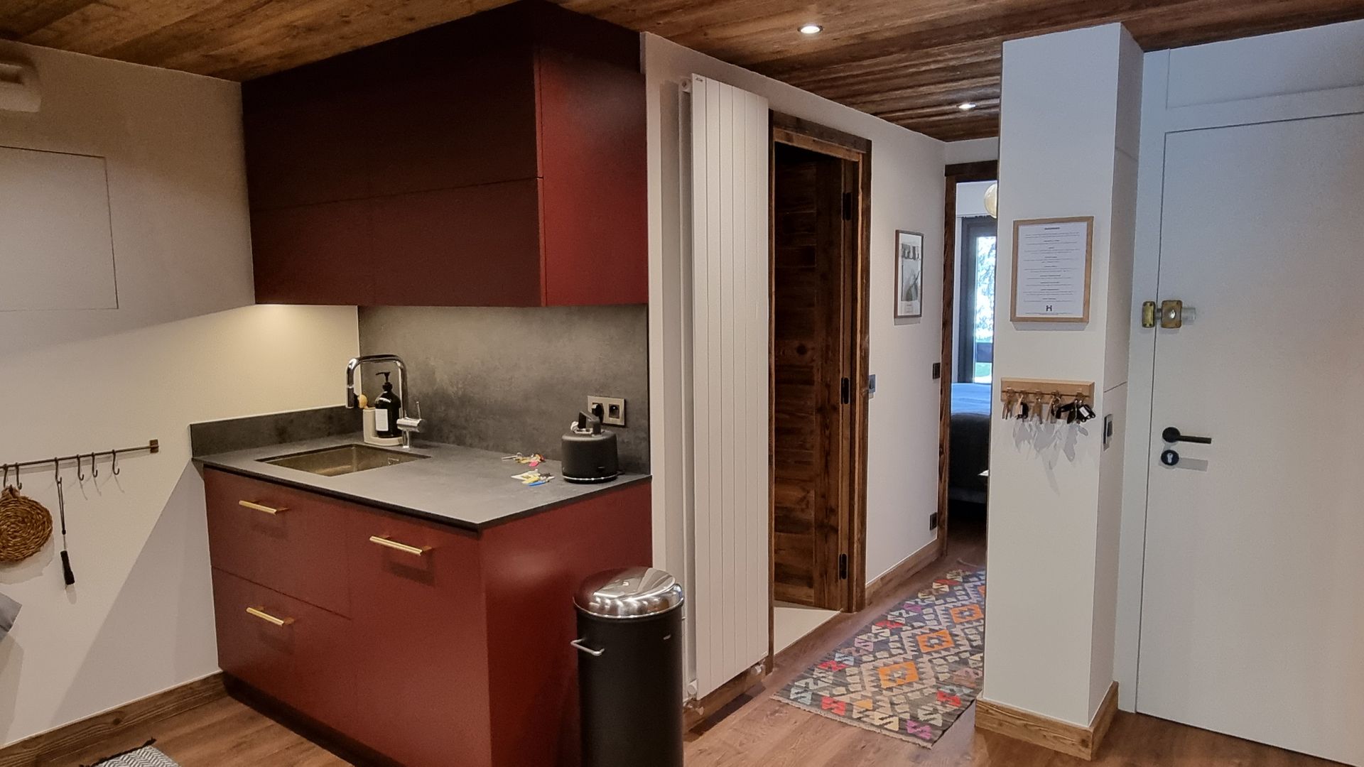 Petite cuisine avec placards rouges, plan de travail en pierre et plafond en bois, donnant sur un couloir.