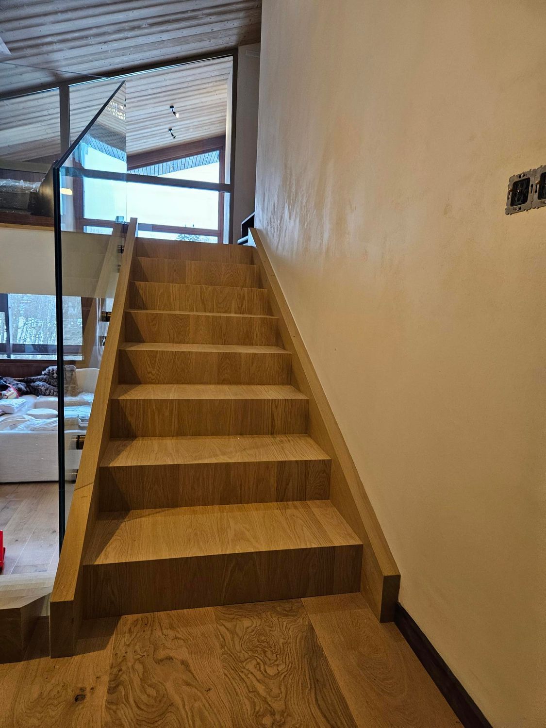Escalier en bois menant à l'étage, avec une rambarde en verre à gauche et un mur de couleur neutre à droite.