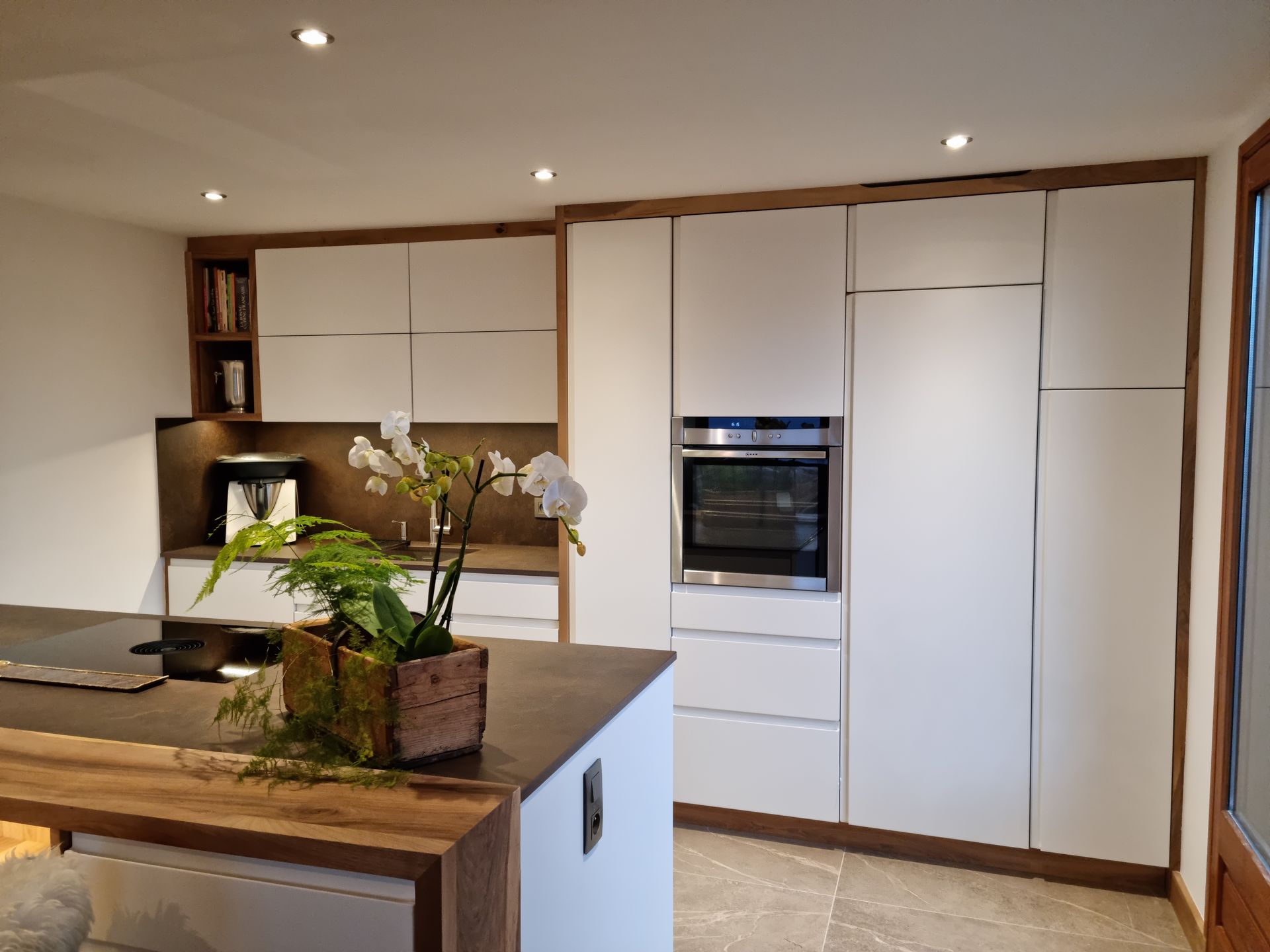 Cuisine moderne blanche avec des touches de bois, un îlot central, un four encastré et une plante.