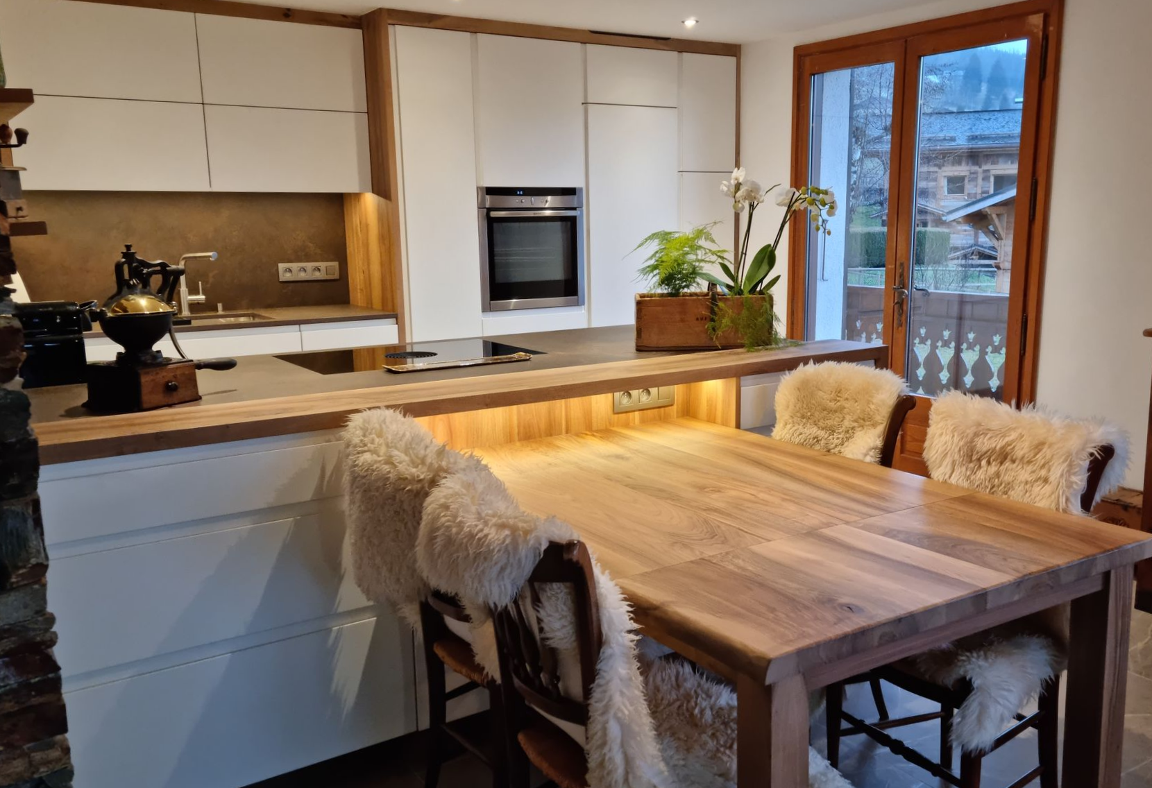 Cuisine moderne avec des éléments en bois et une table à manger, comprenant des armoires blanches, un four encastré et une vue sur un paysage de montagne.