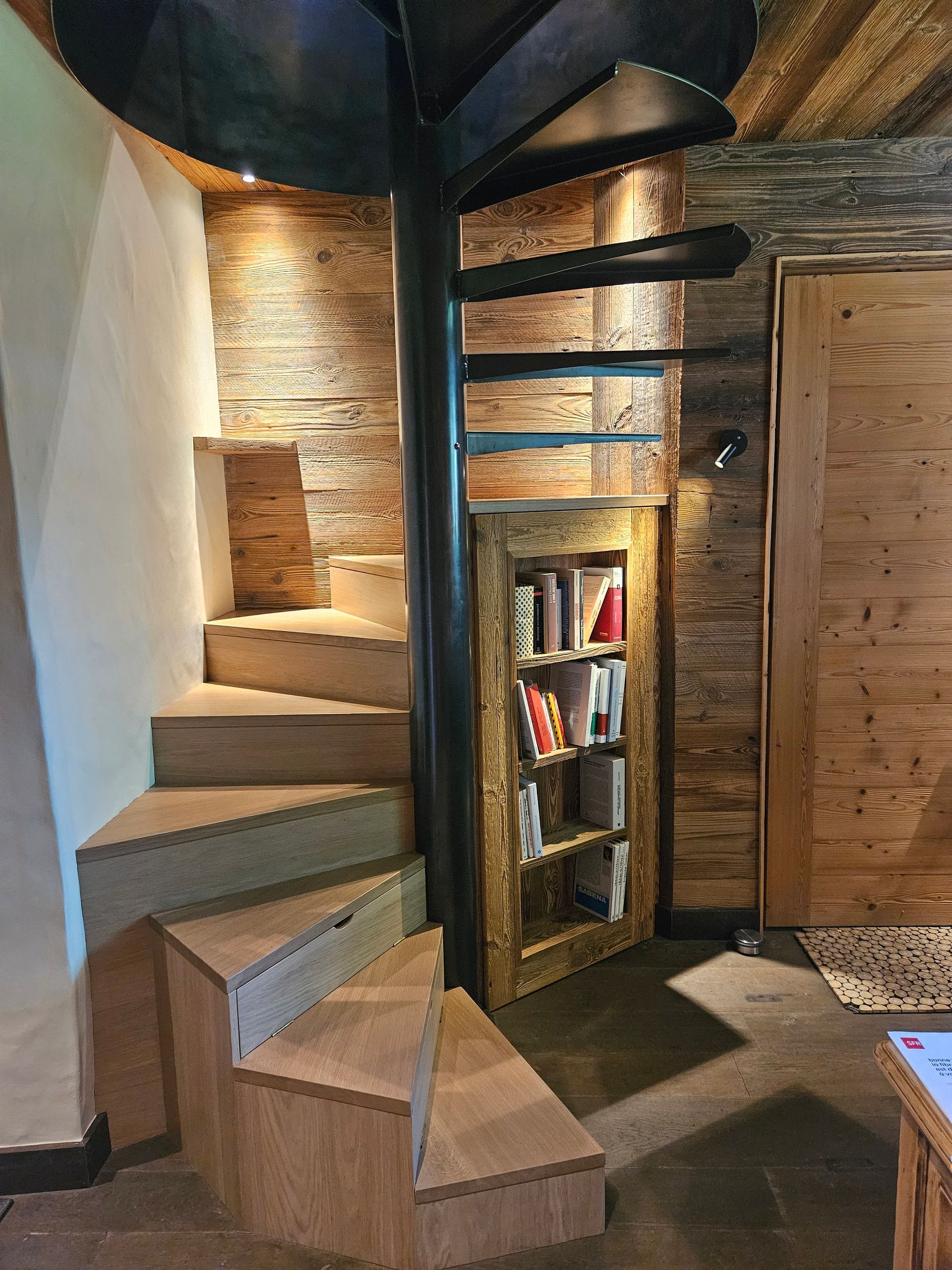 Escalier en colimaçon avec marches en bois et pilier central en métal noir, avec bibliothèque intégrée.