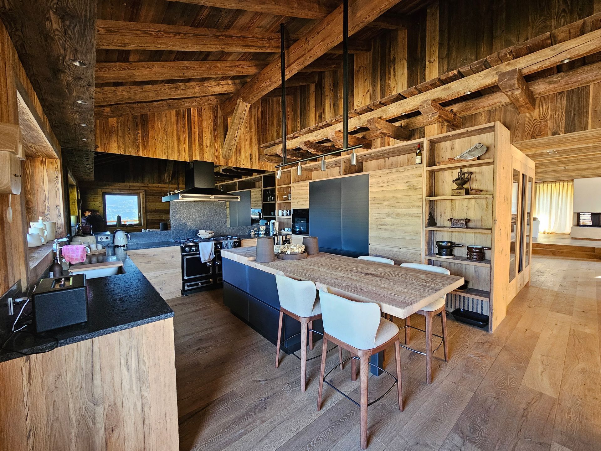 Cuisine rustique avec poutres et îlot en bois, comptoirs foncés et chaises blanches.