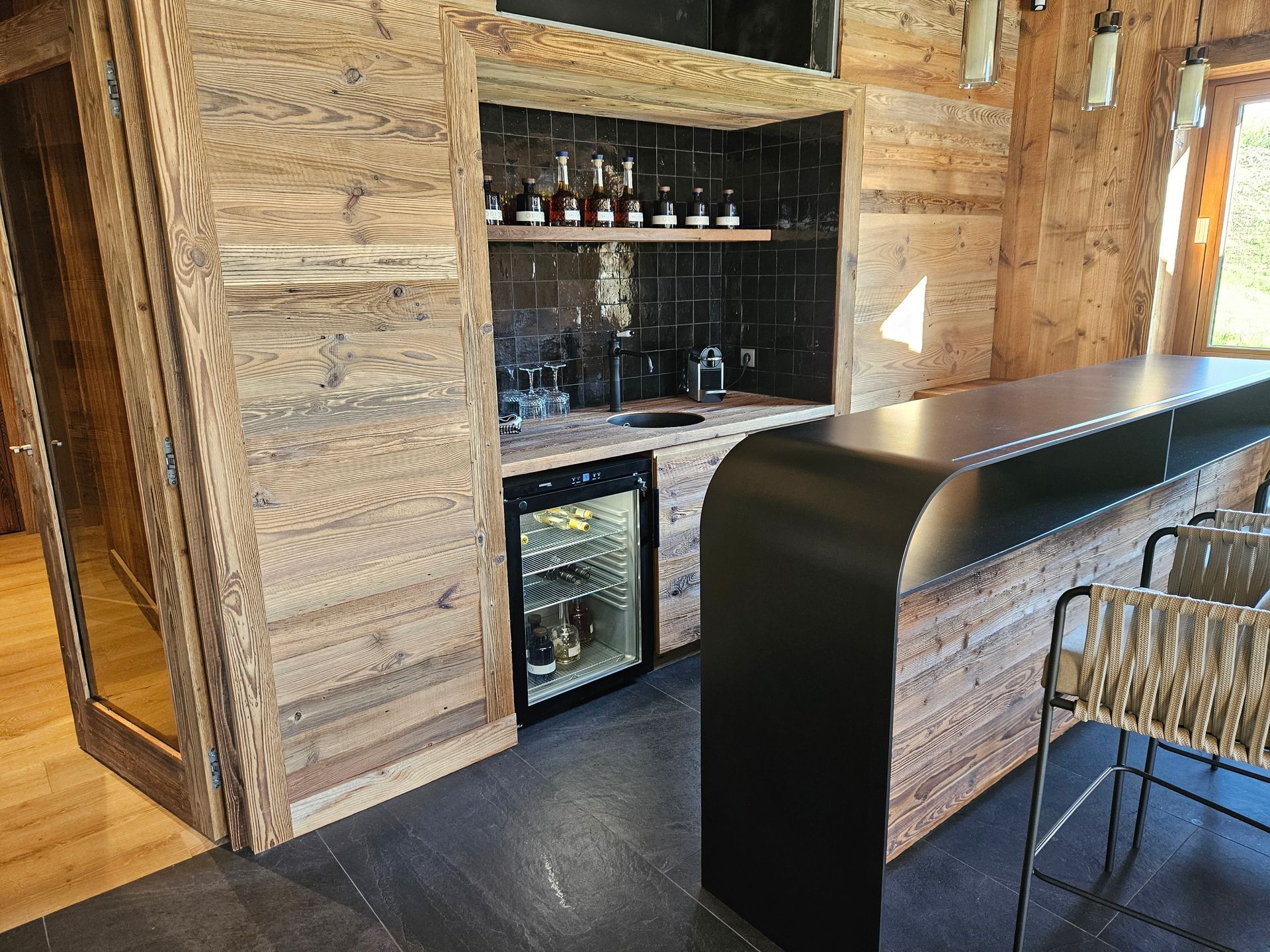 Bar rustique avec murs en bois, comptoir noir, mini-réfrigérateur intégré et sièges.