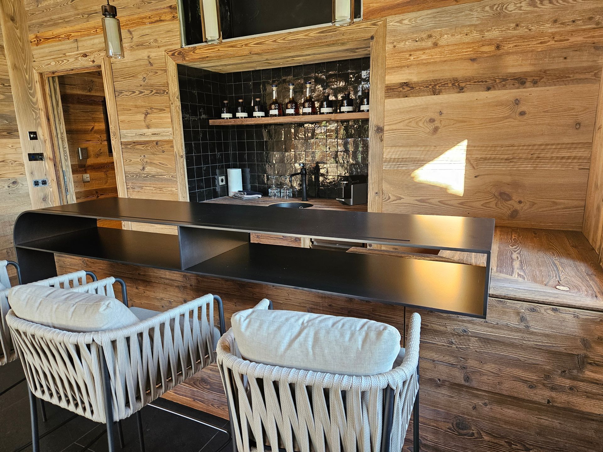 Espace bar moderne avec comptoir noir, boiseries, bouteilles et deux chaises en corde blanche.