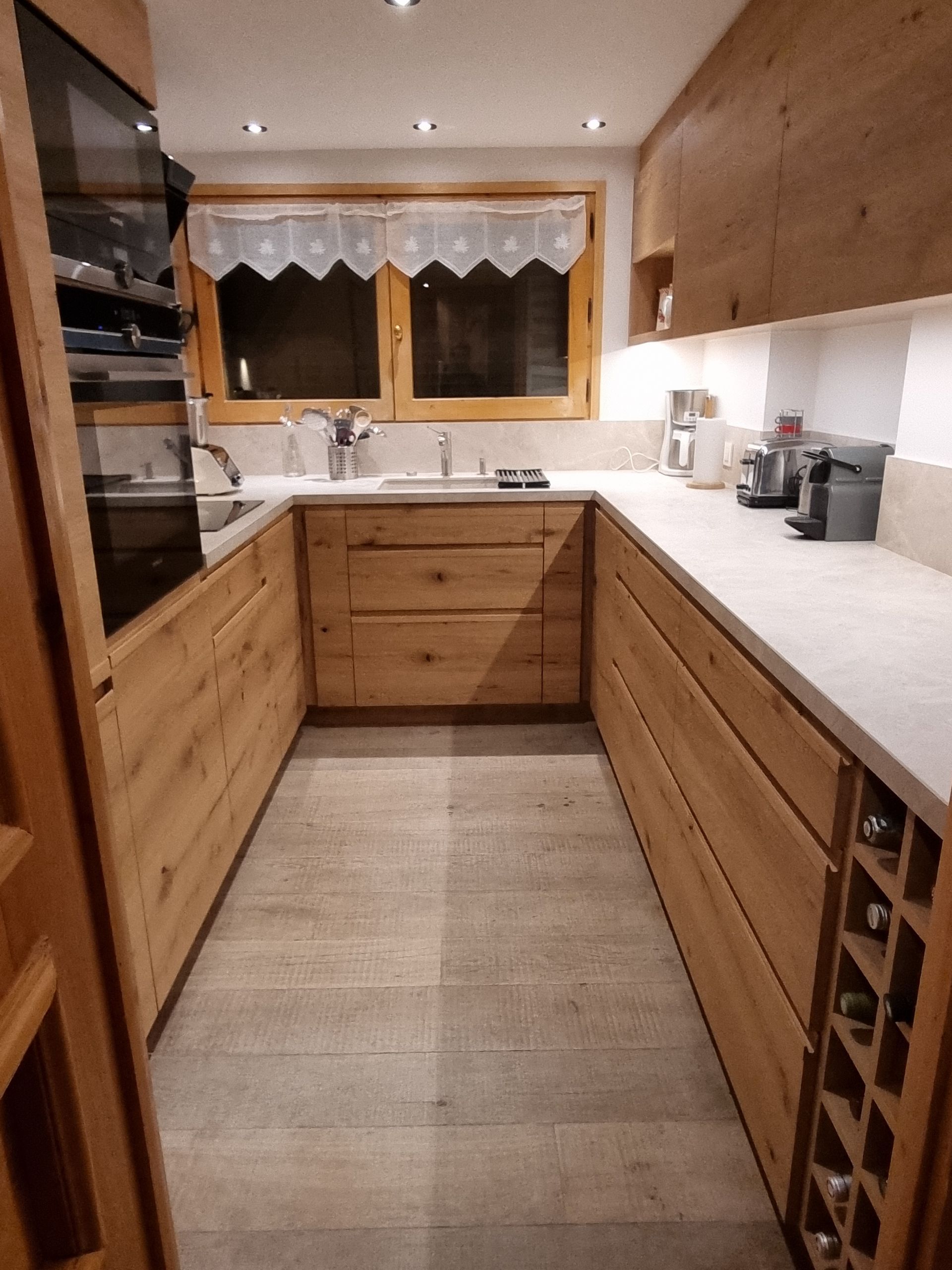 Cuisine en forme de U avec placards en bois, plan de travail clair, fenêtre avec rideau, électroménager intégré et sol carrelé.