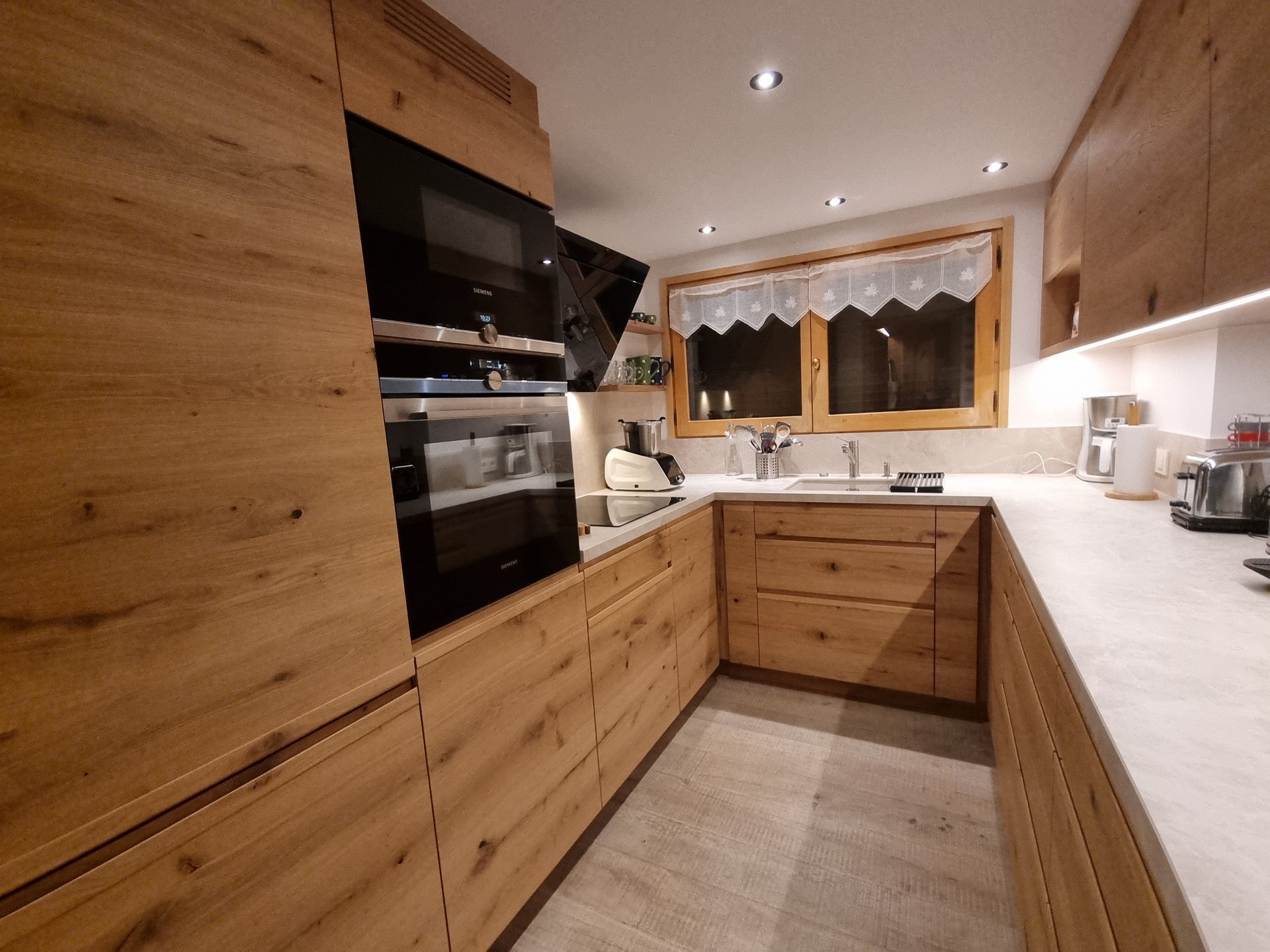 Cuisine en bois avec électroménager intégré, tiroirs et plan de travail. Une fenêtre et des luminaires sont visibles.