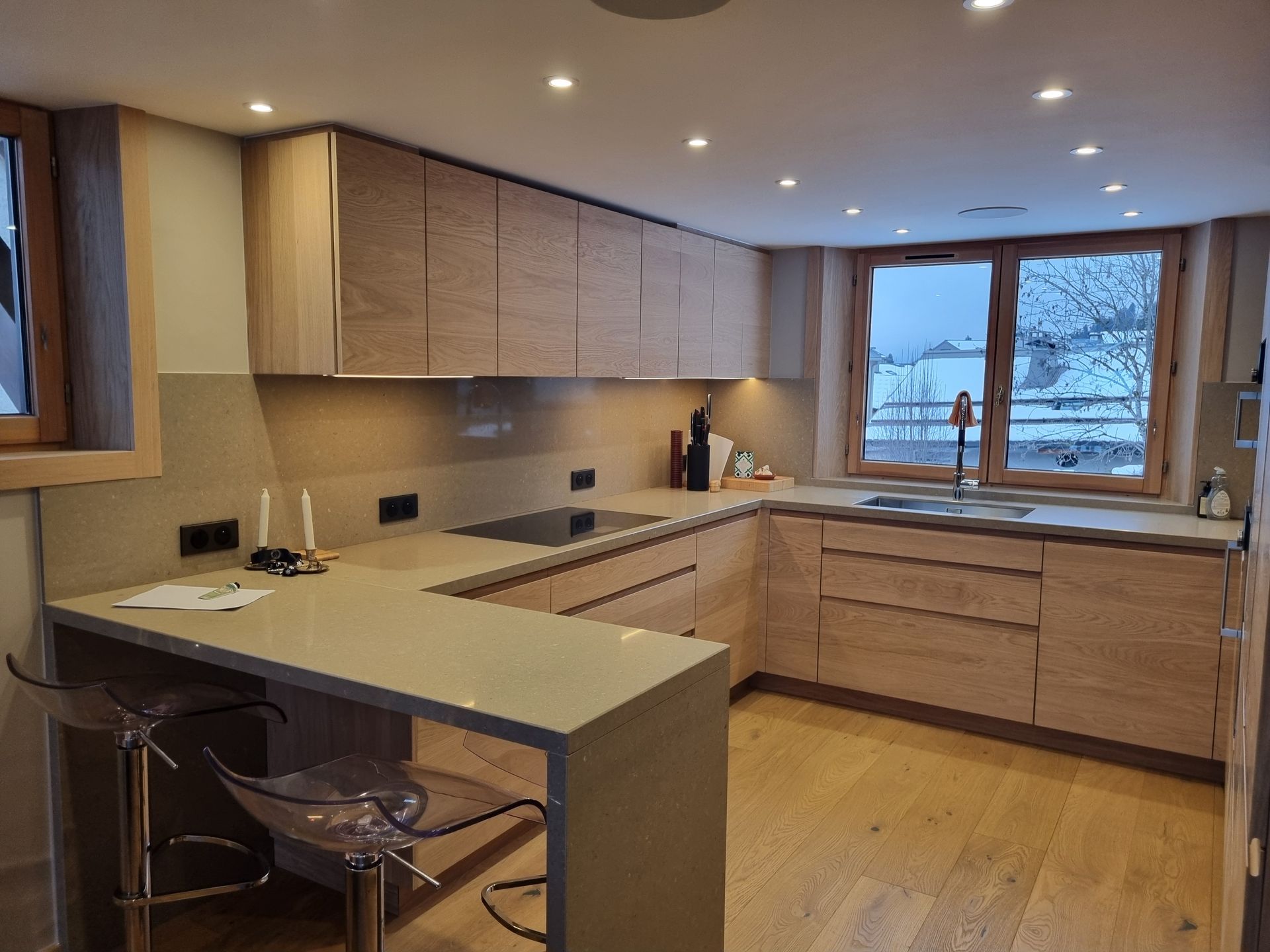 Cuisine moderne avec placards et plans de travail en bois clair, un bar pour le petit-déjeuner et une vue sur un paysage enneigé.