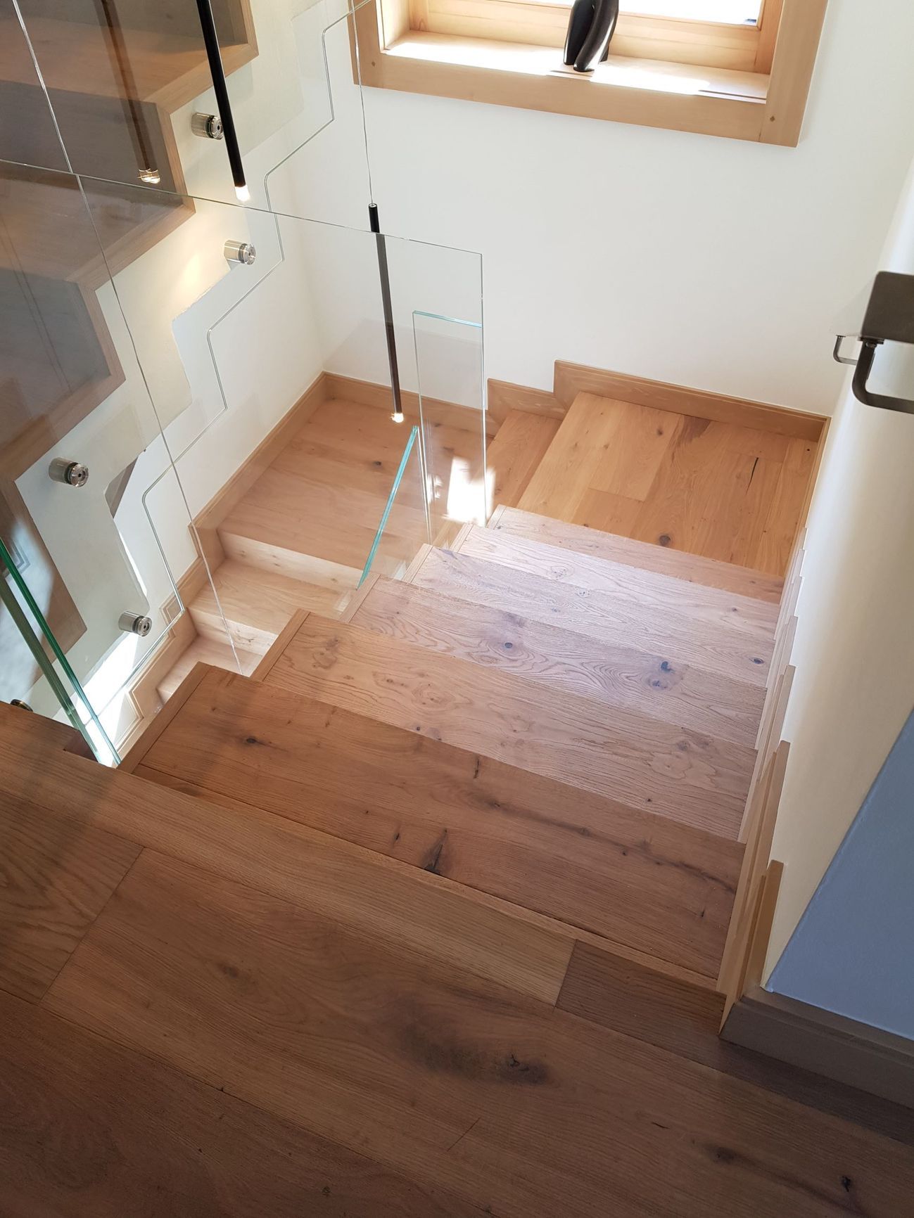 Escalier en bois avec rambarde en verre, vu d'en haut, dans une pièce lumineuse.