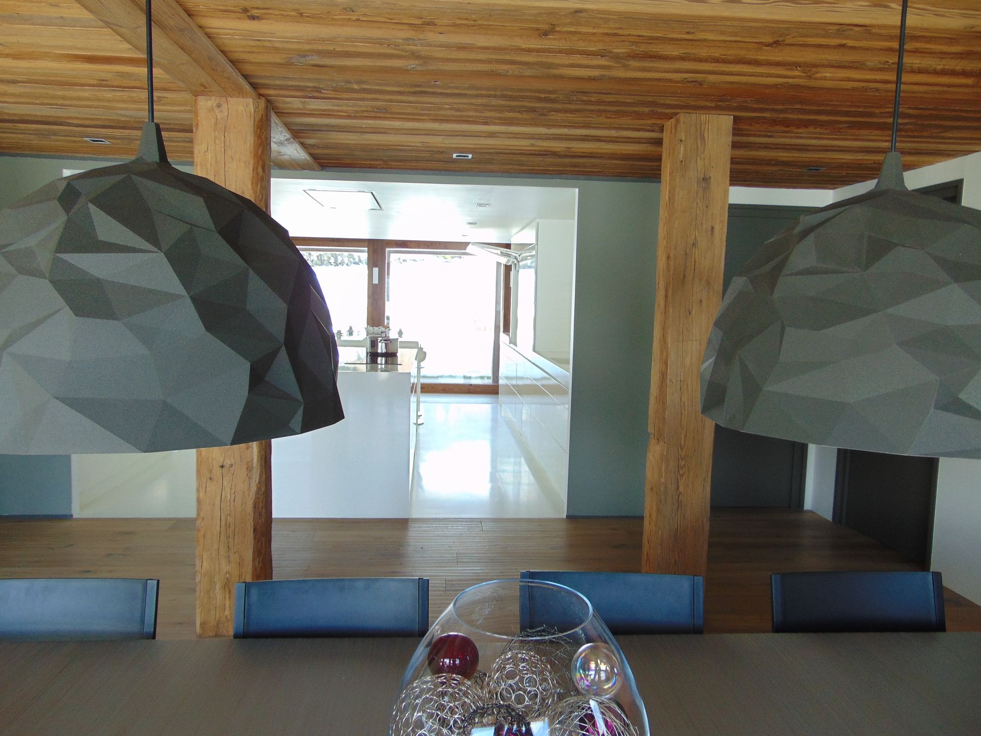 Salle à manger avec lampes suspendues, poutres en bois, table, chaises et vue sur une autre pièce.