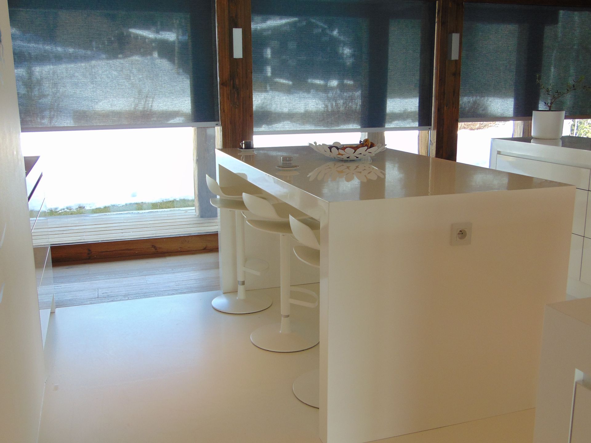 Îlot de cuisine blanc avec trois tabourets, à côté d'une fenêtre avec stores occultants.