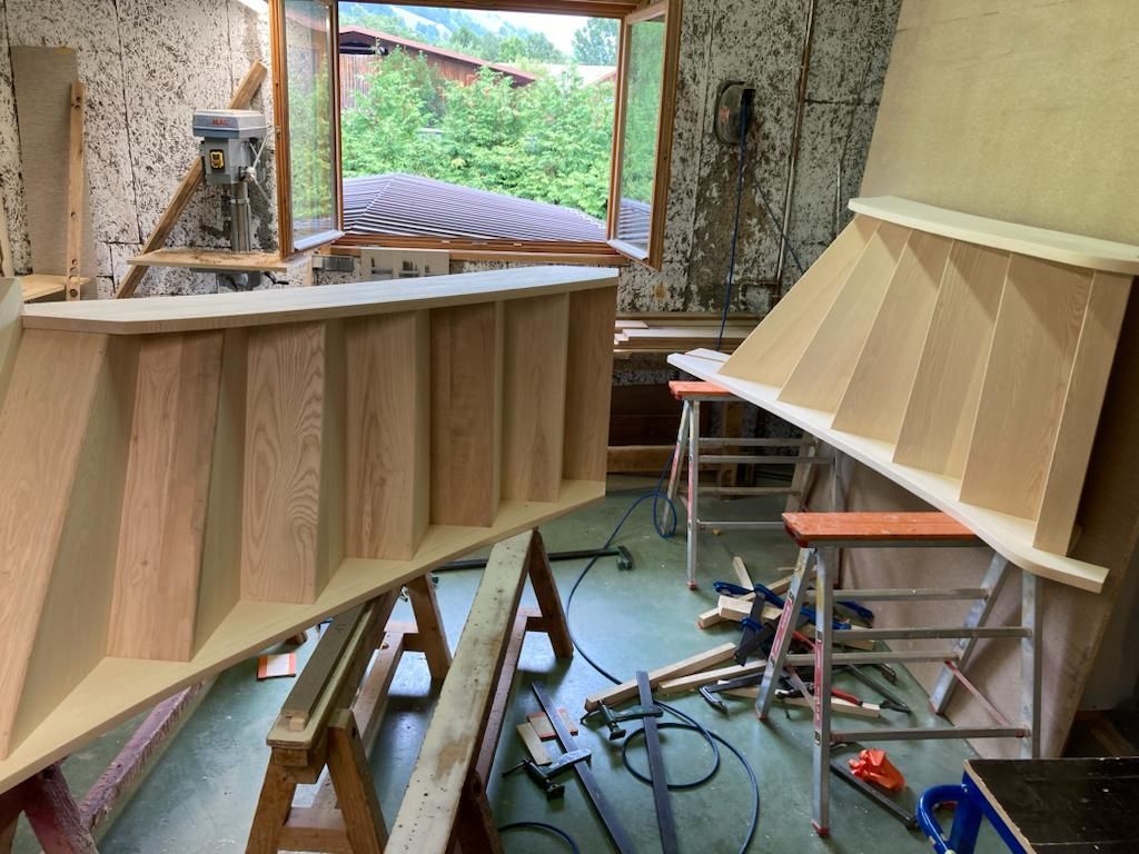 Éléments d'escalier en bois dans un atelier en construction. L'un est assemblé, l'autre partiellement construit, près d'une fenêtre.