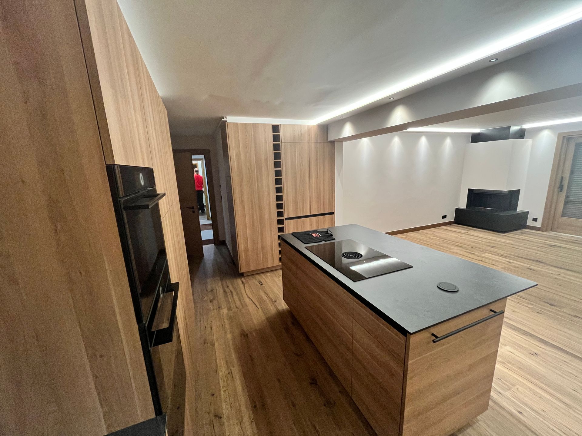 Cuisine moderne avec placards en bois, îlot central et électroménager. Une personne est visible dans le couloir.