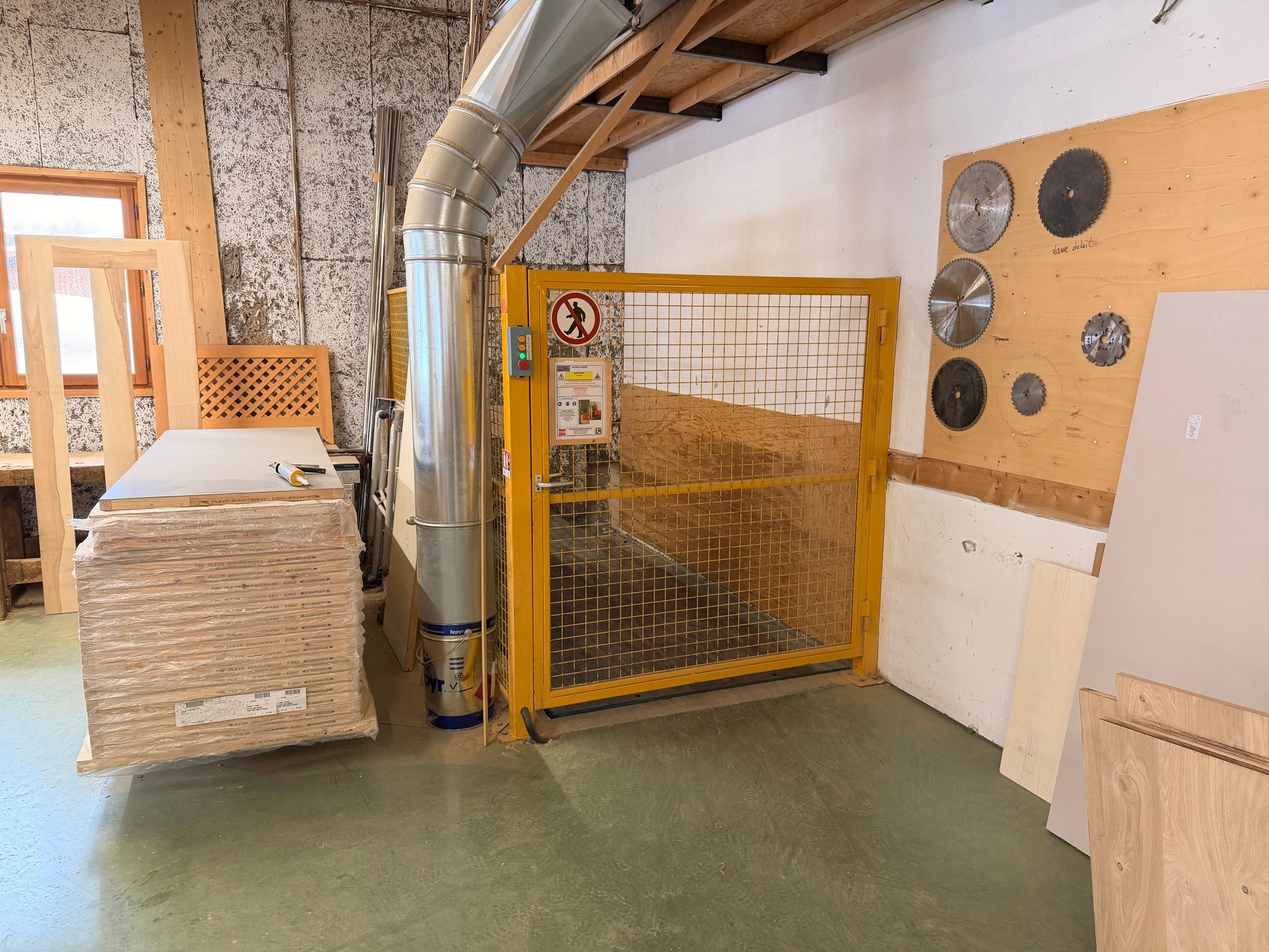 Intérieur d'atelier avec porte de sécurité jaune, système d'aspiration des poussières, piles de bois et lames de coupe.