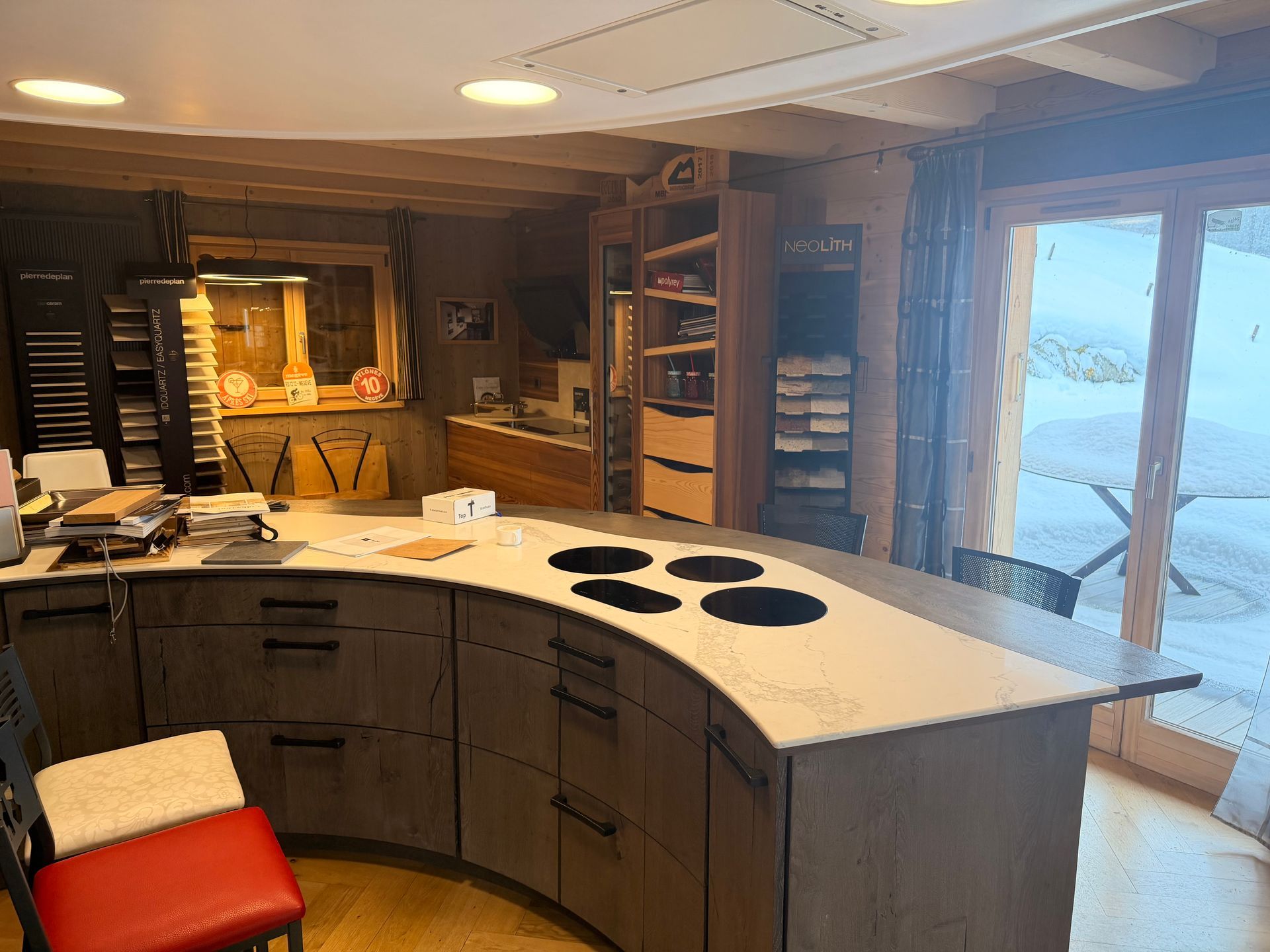 Cuisine avec îlot central incurvé, plaque de cuisson intégrée, placards en bois et vue sur un paysage enneigé.