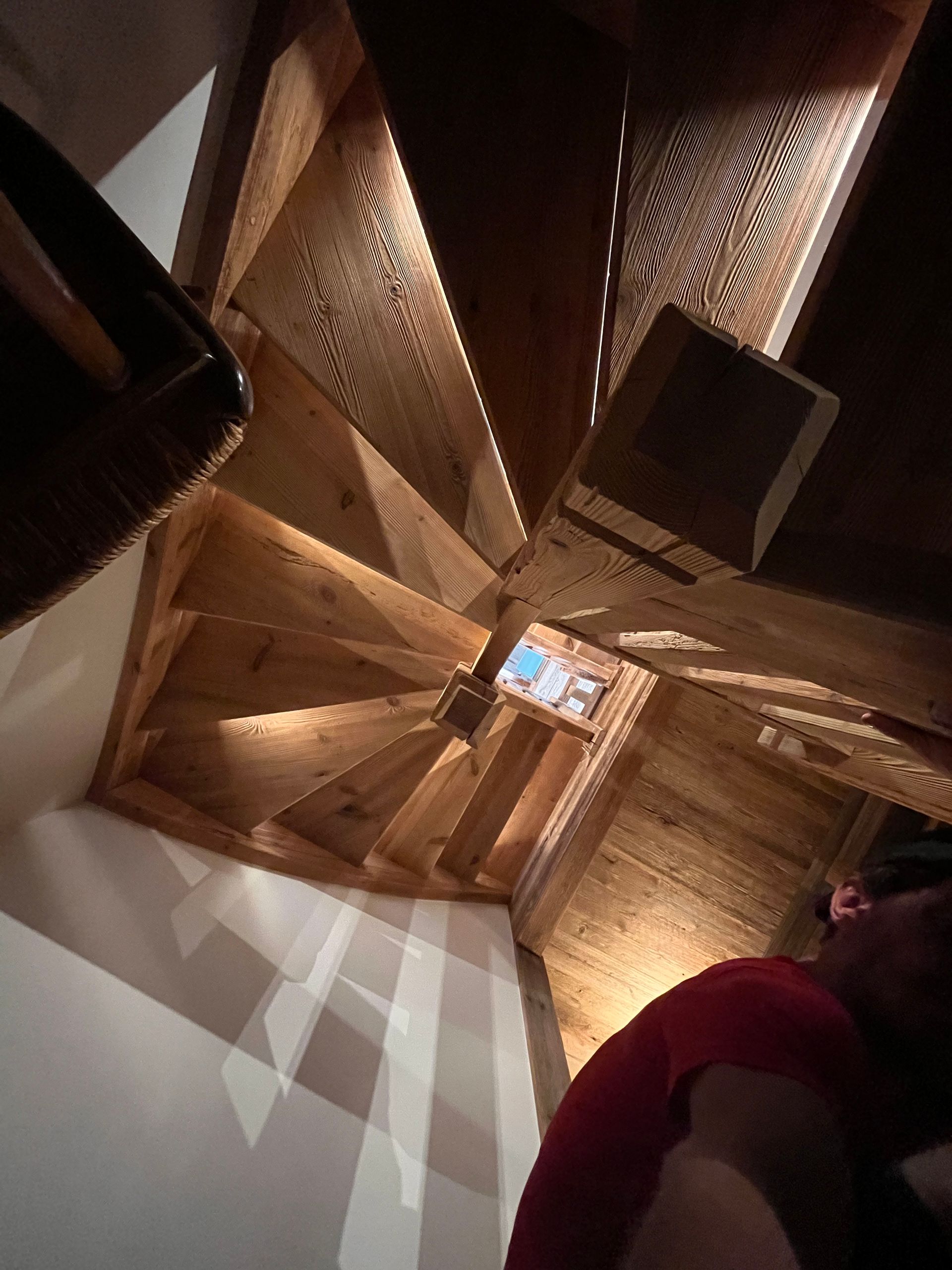 Escalier en colimaçon en bois éclairé par le haut, personne en chemise rouge en bas.