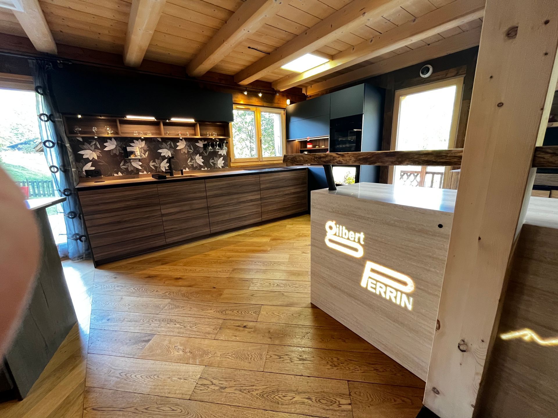 Cuisine à dominante bois avec îlot central ; logo Burt & Perrin sur l’îlot. Sol, placards et plafond en bois.