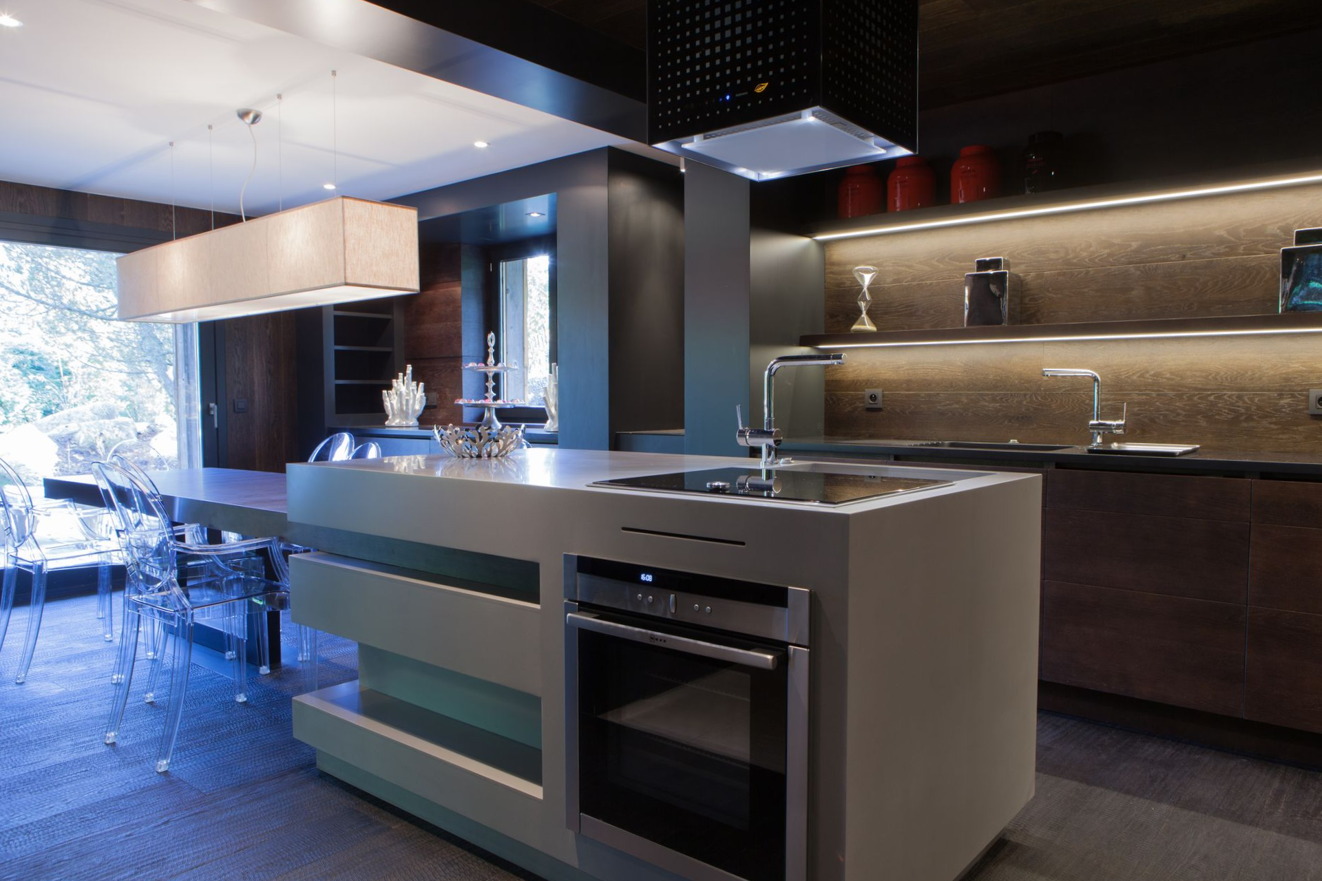 Cuisine moderne avec îlot central, four et évier gris. Placards en bois foncé avec éclairage sous les meubles.