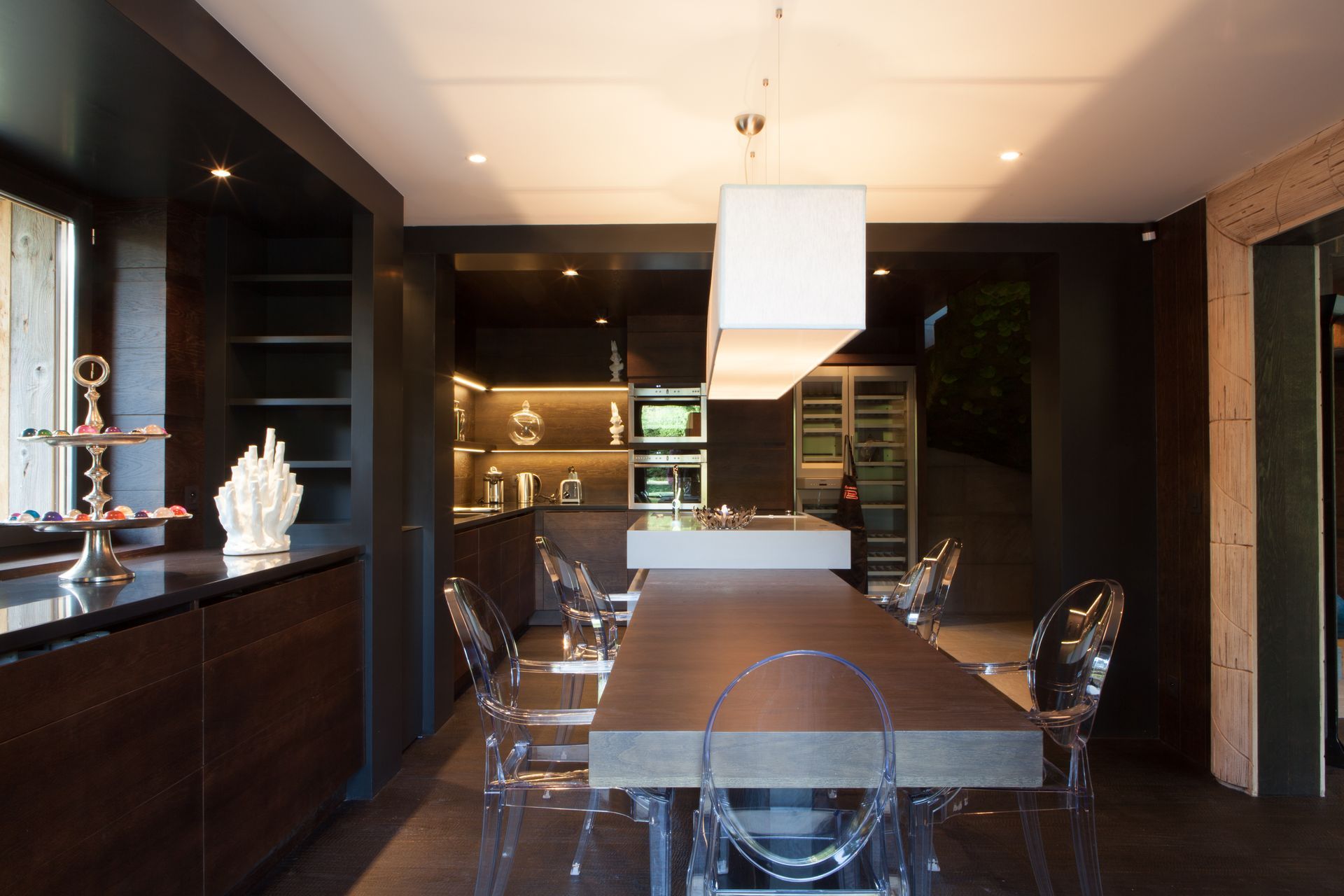 Salle à manger moderne avec une longue table, des chaises transparentes, des meubles de cuisine foncés et une grande suspension lumineuse.