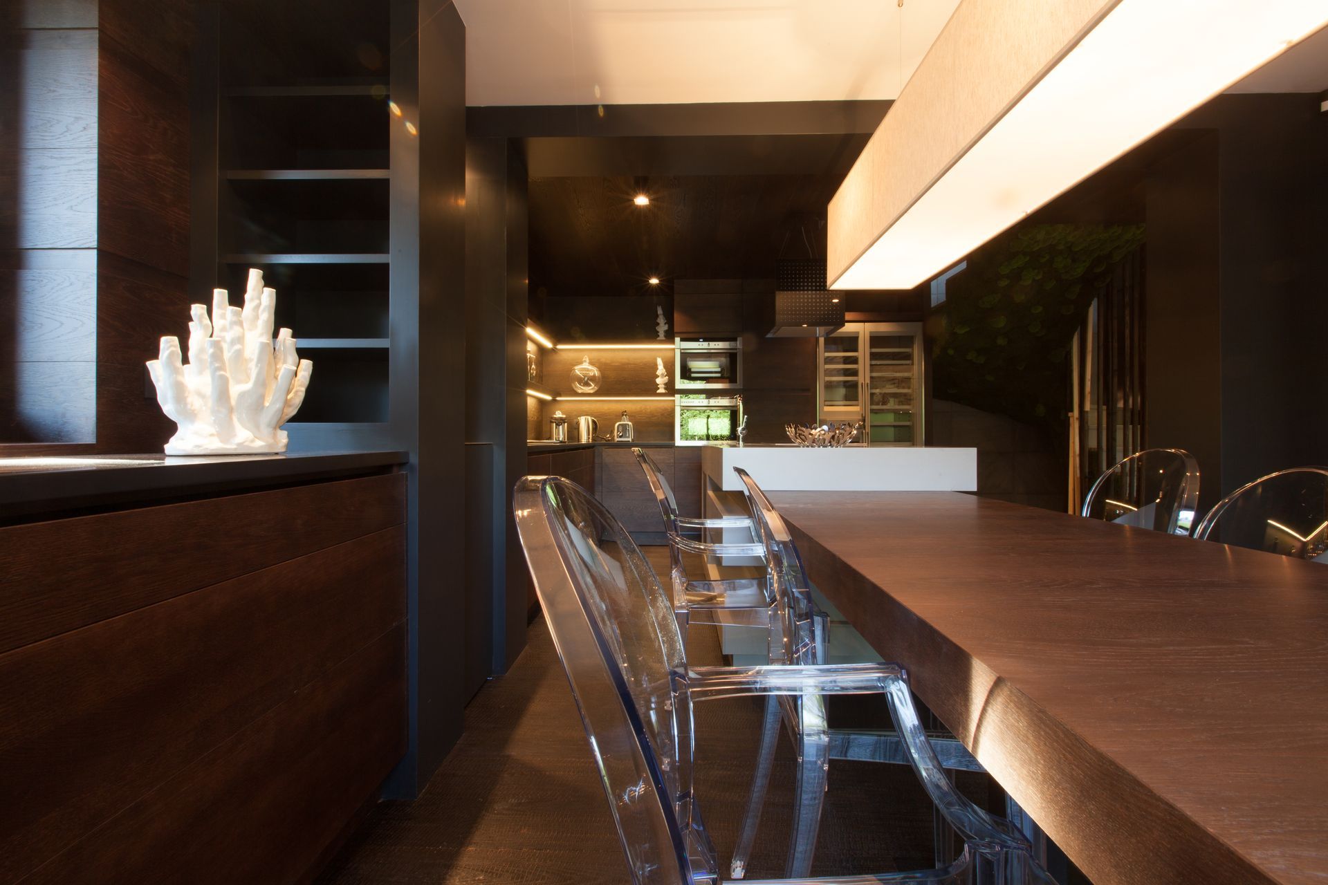Intérieur de cuisine moderne avec bois foncé et chaises transparentes, éclairage vif.