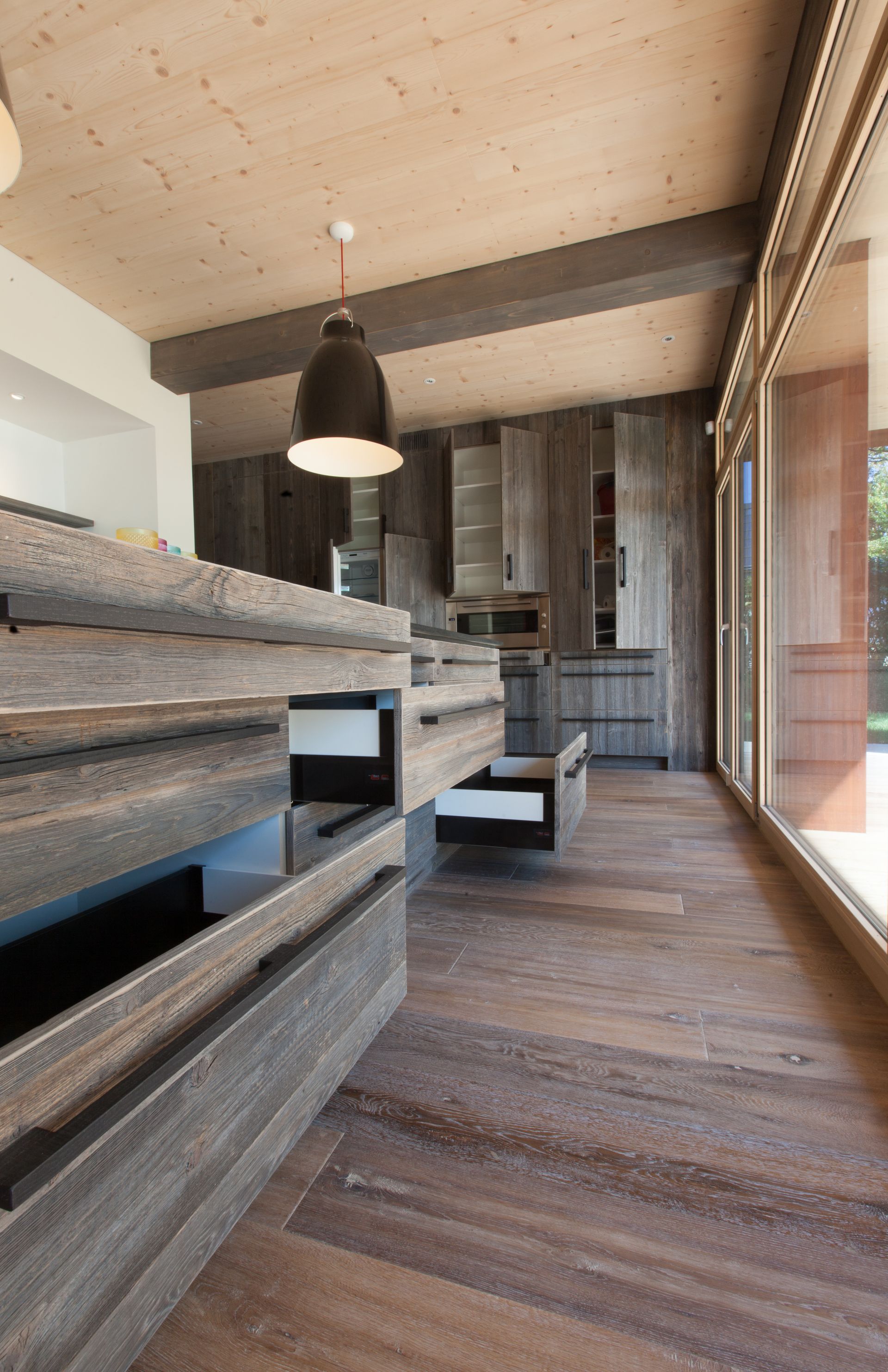 Cuisine en bois avec tiroirs ouverts, luminaire, plafond en bois et grande fenêtre.