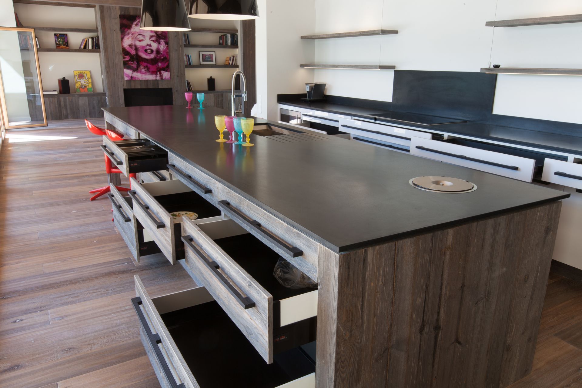 Îlot de cuisine avec tiroirs ouverts, plan de travail foncé, placards en bois et tasses colorées.