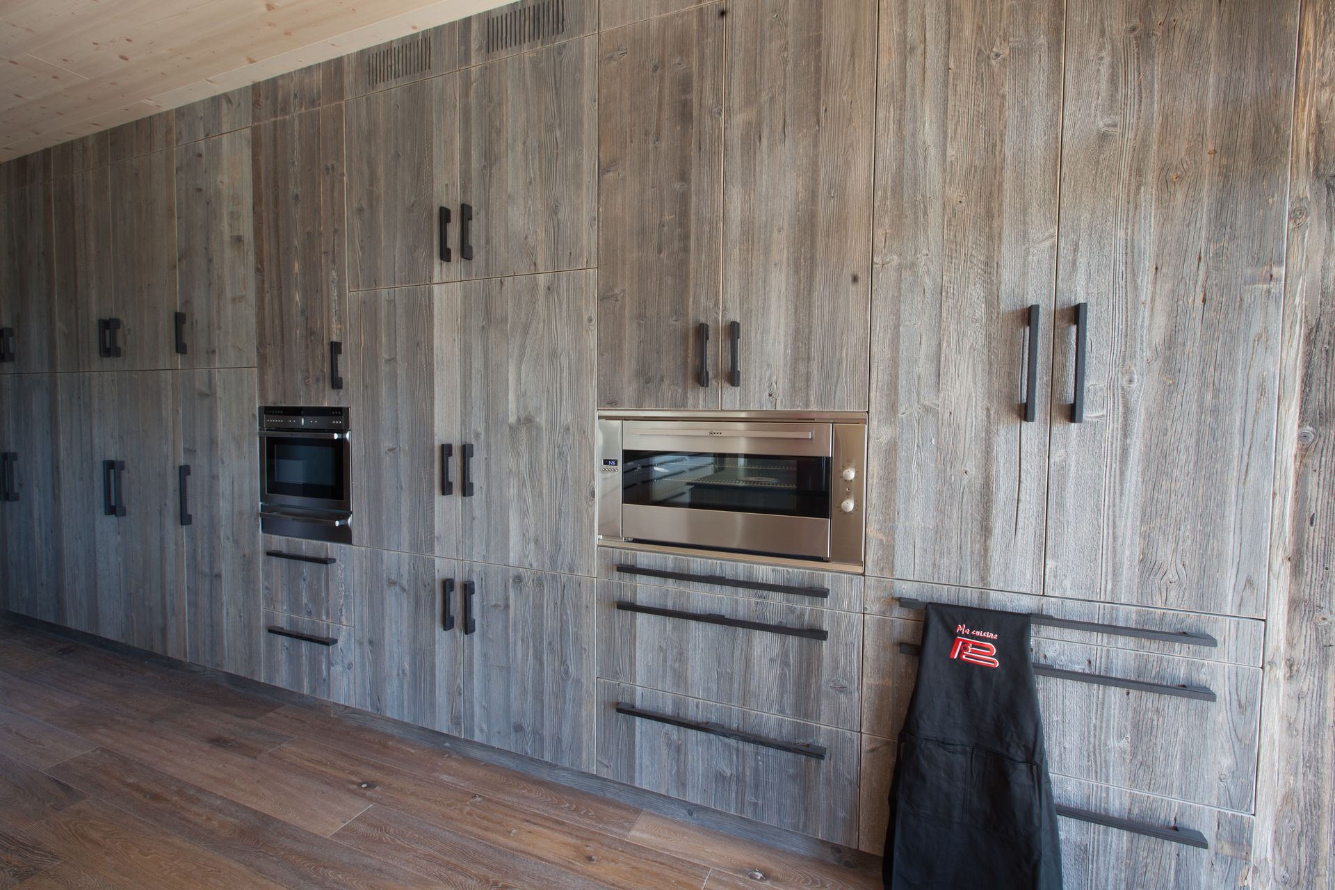 Cuisine équipée d'armoires en bois gris avec four et micro-ondes intégrés. Quincaillerie noire, tablier suspendu, parquet.