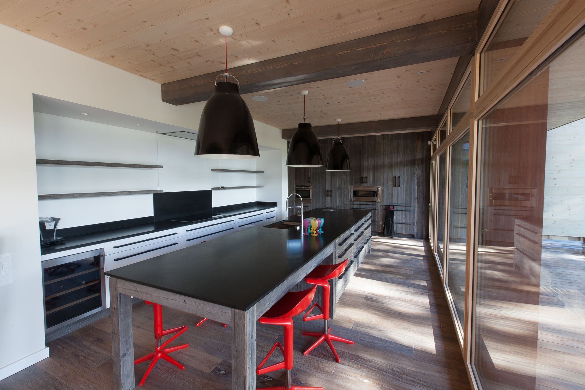 Cuisine moderne avec îlot central noir, tabourets rouges, étagères ouvertes et grandes fenêtres.