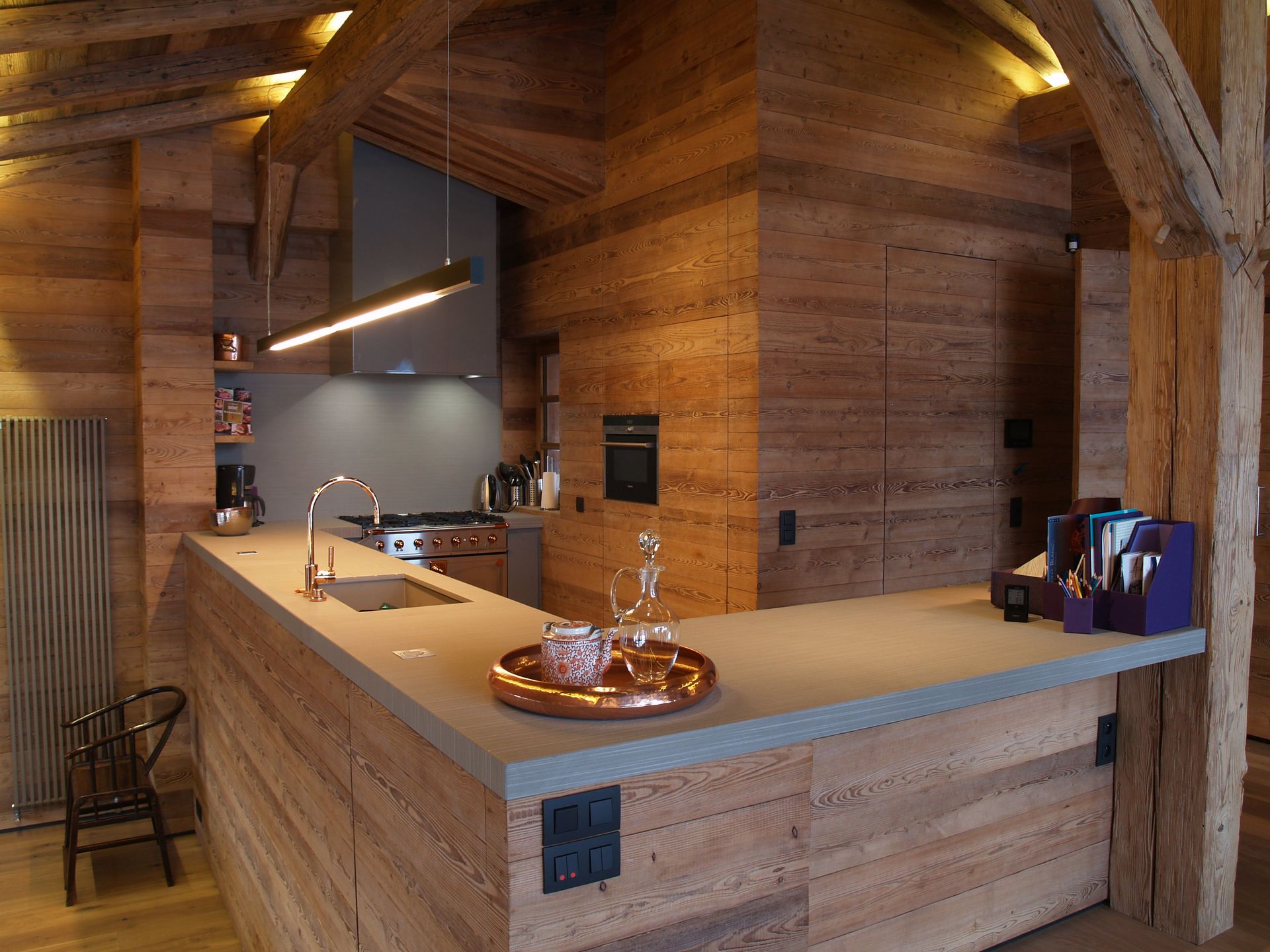 Cuisine avec murs en pisé, poutres en bois, plans de travail gris et appareils électroménagers modernes.