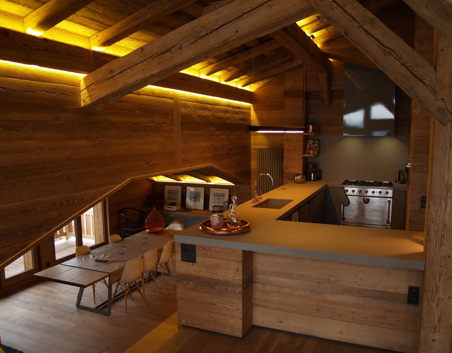 Cuisine en bois chaleureuse avec bar, table à manger et poutres apparentes, éclairée par une lumière chaude.