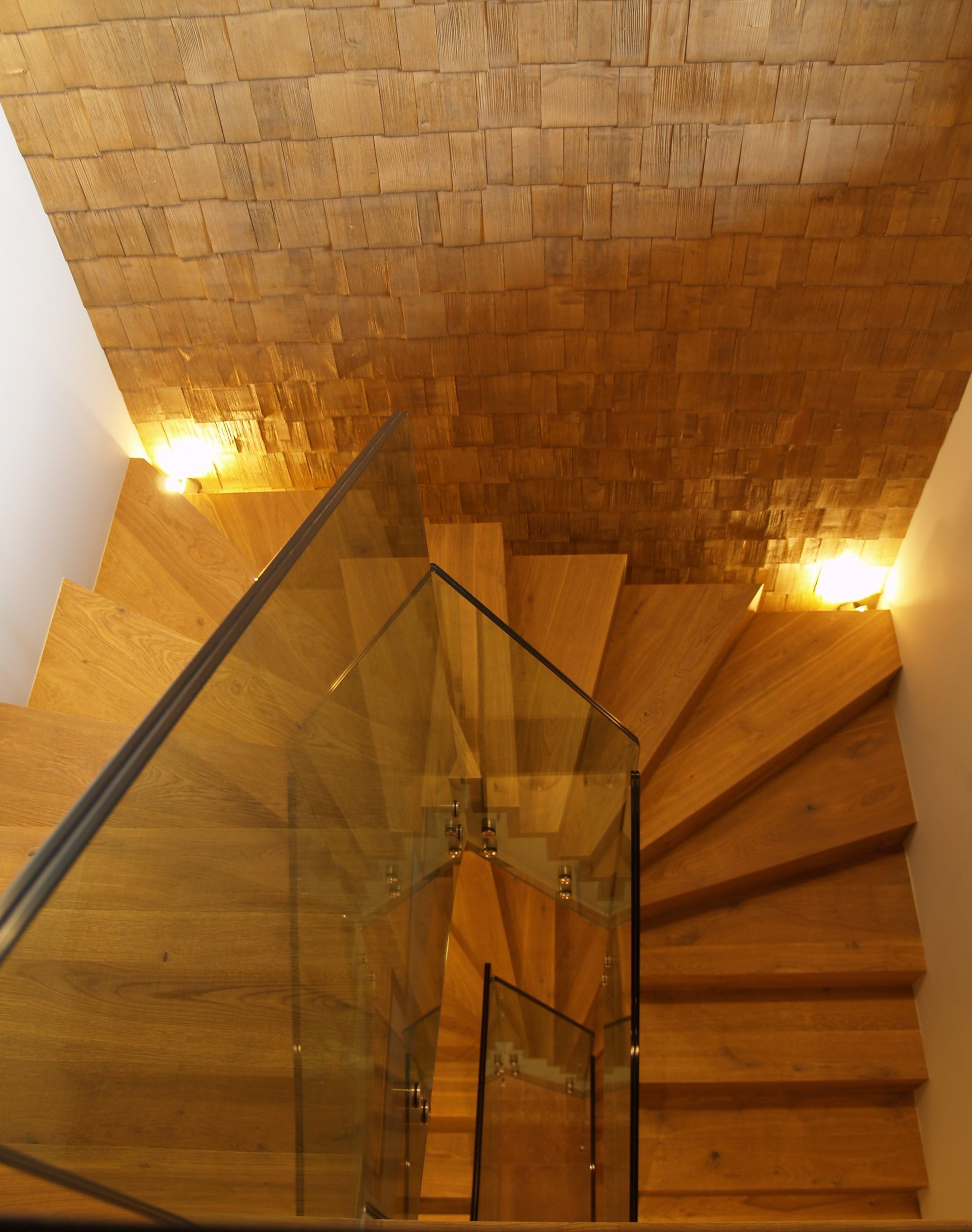 Escalier en bois avec rampe en verre ; éclairage chaleureux par appliques murales ; vue d'en haut.