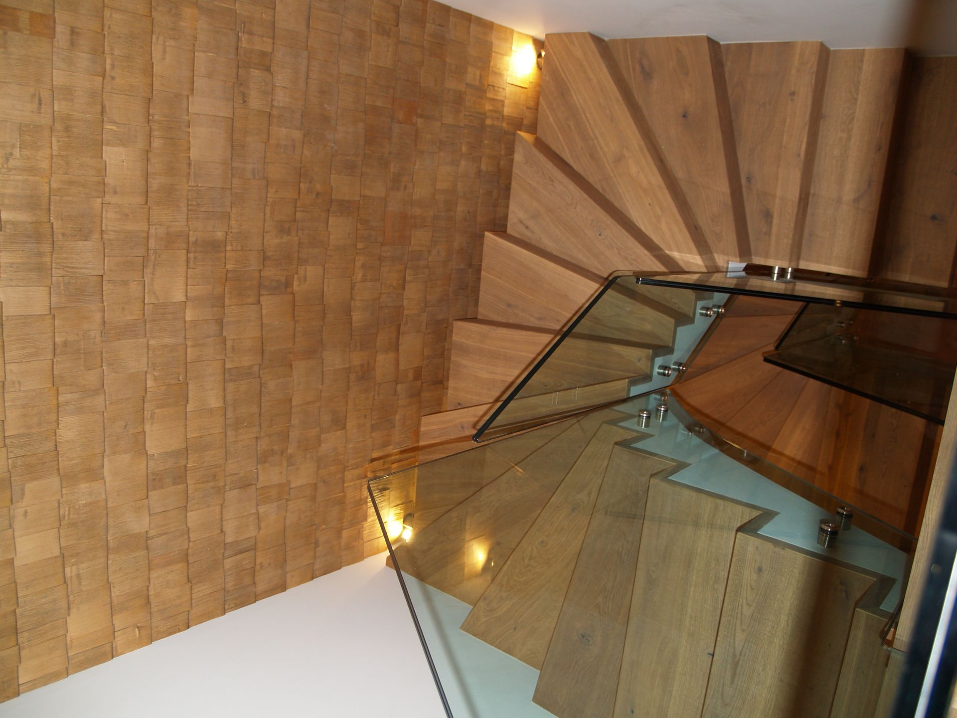 Escalier en colimaçon en bois avec rampe en verre et mur texturé.