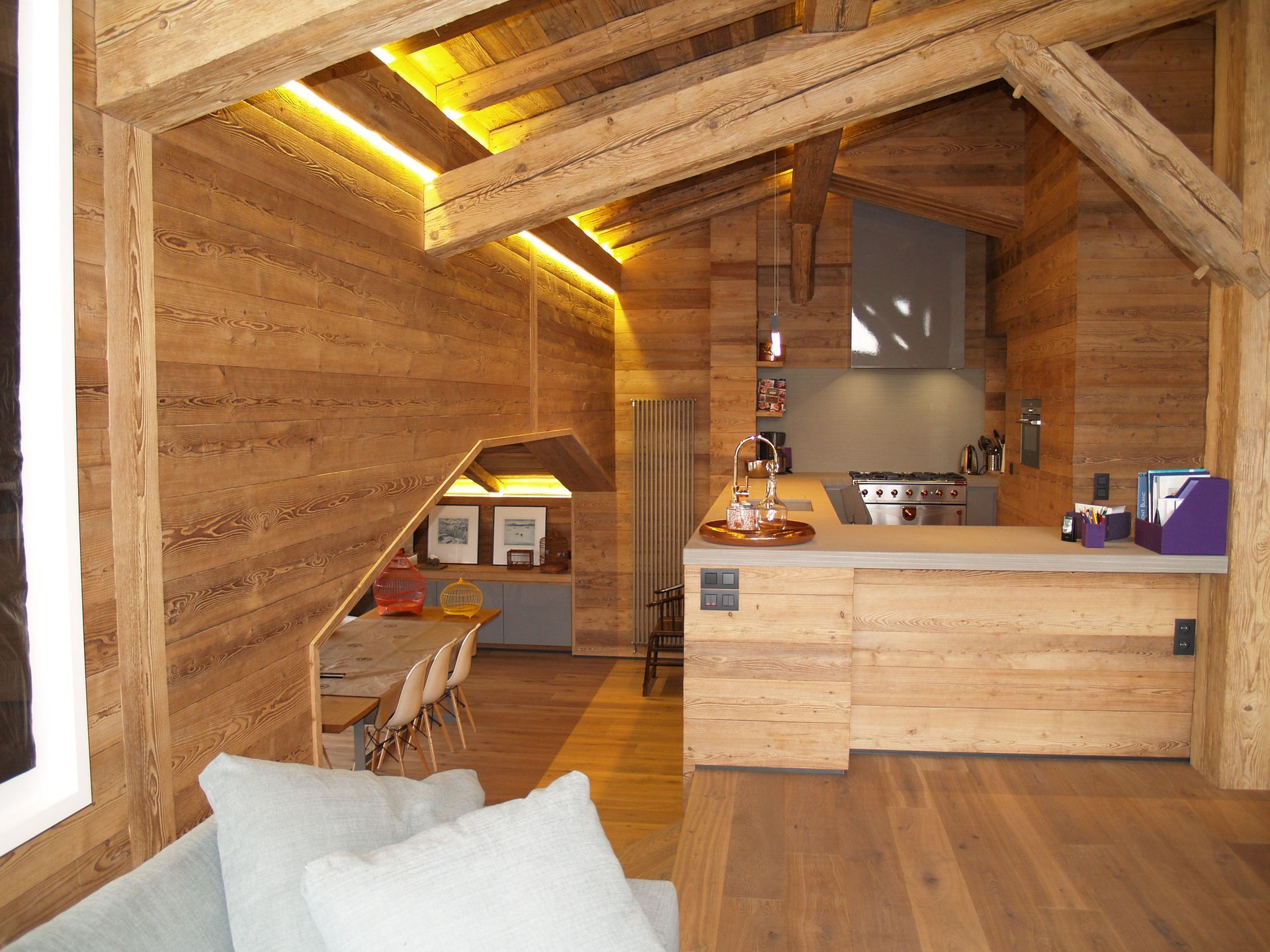 Intérieur chaleureux en bois avec cuisine, coin repas et salon. Un éclairage doux et des poutres apparentes créent une ambiance conviviale.