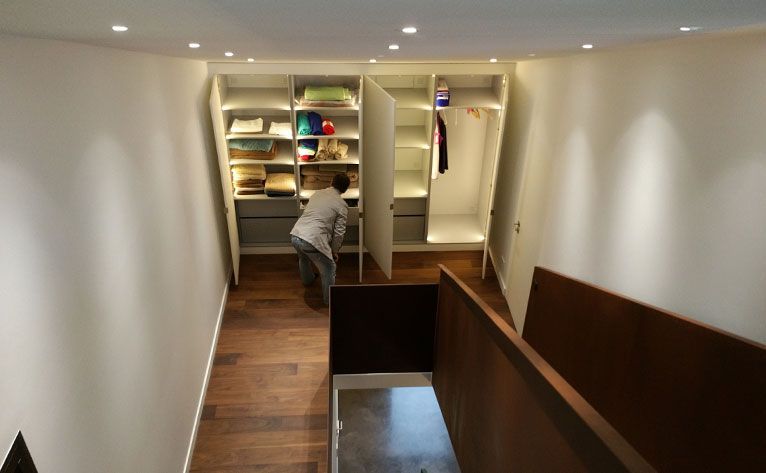 Une personne range ses vêtements dans un placard blanc éclairé par des spots encastrés, intégré dans un mur incliné.